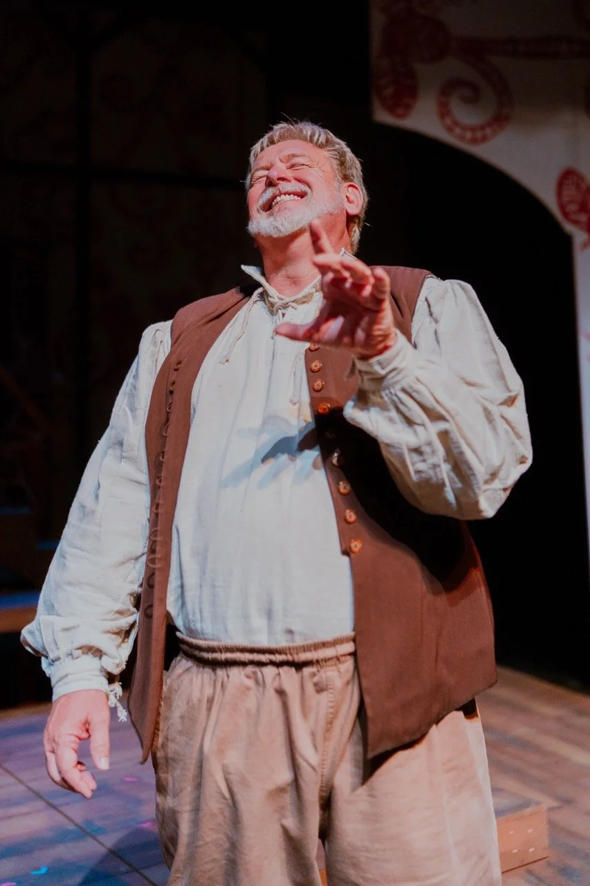 A person in historical costume, smiling, wearing a white shirt with a brown vest and beige pants, performing on stage.
