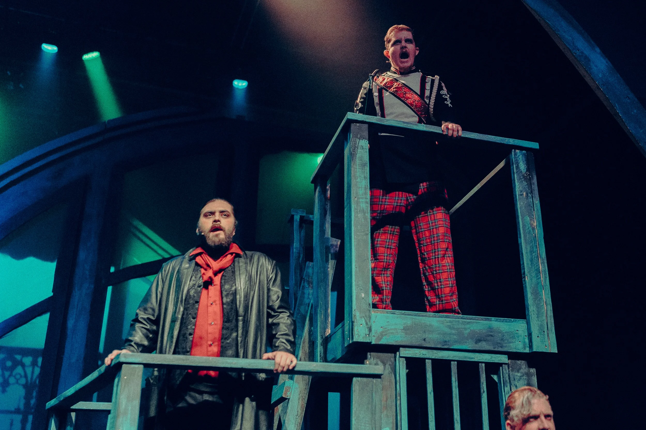 Two actors on stage, one in a black leather jacket and red shirt, the other in a military-style jacket and plaid pants, on a wooden structure with theatrical lighting.