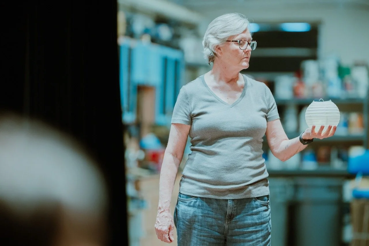 Older woman in glasses holding a white paper lantern indoors.