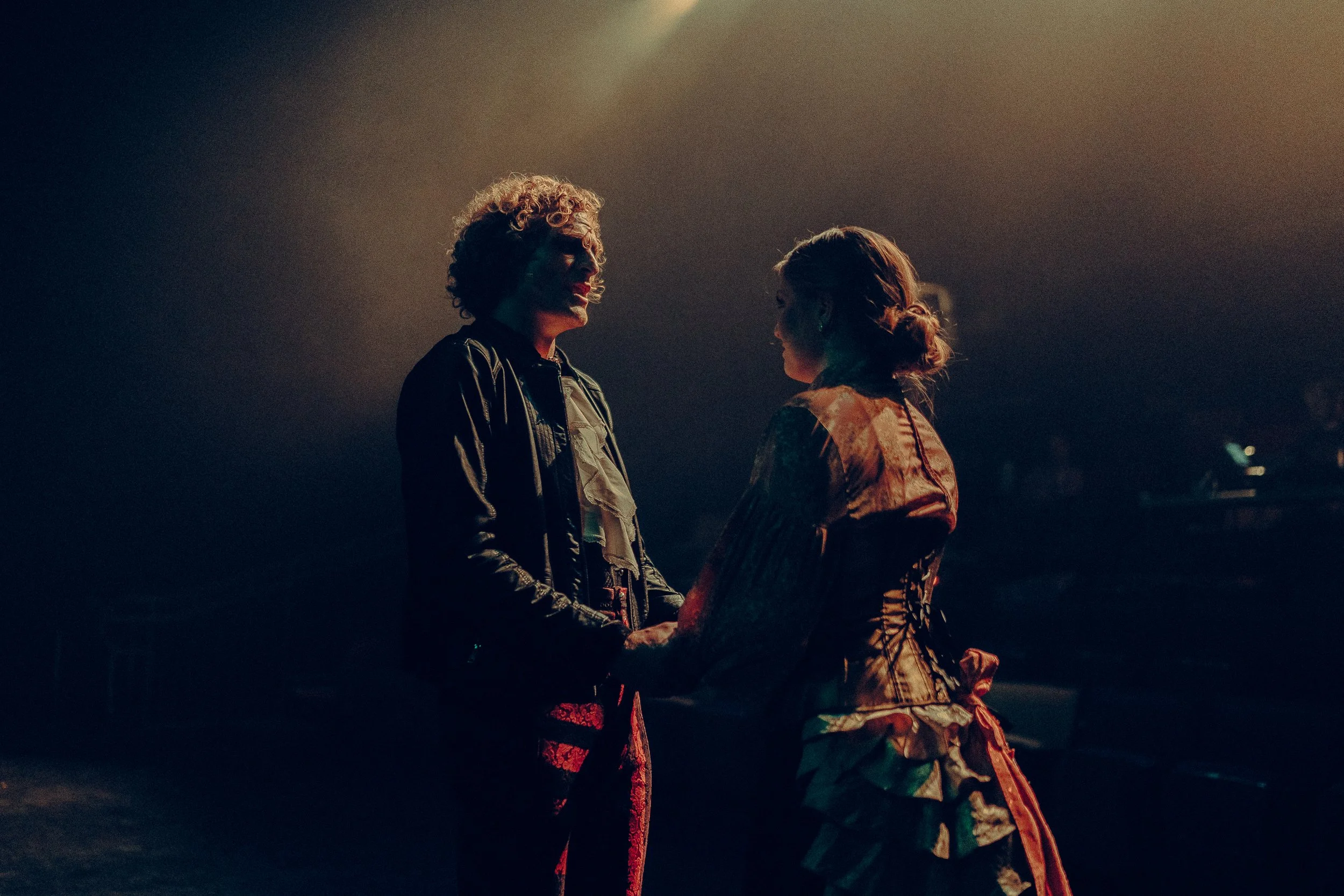 Two people in a dimly lit theater holding hands, wearing vintage-style clothing, with atmospheric stage lighting.