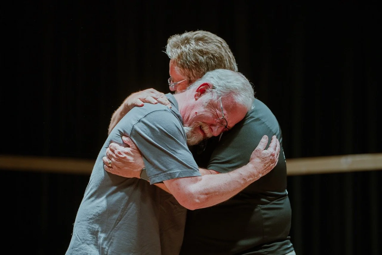 Two men embracing emotionally on a stage with a dark background.