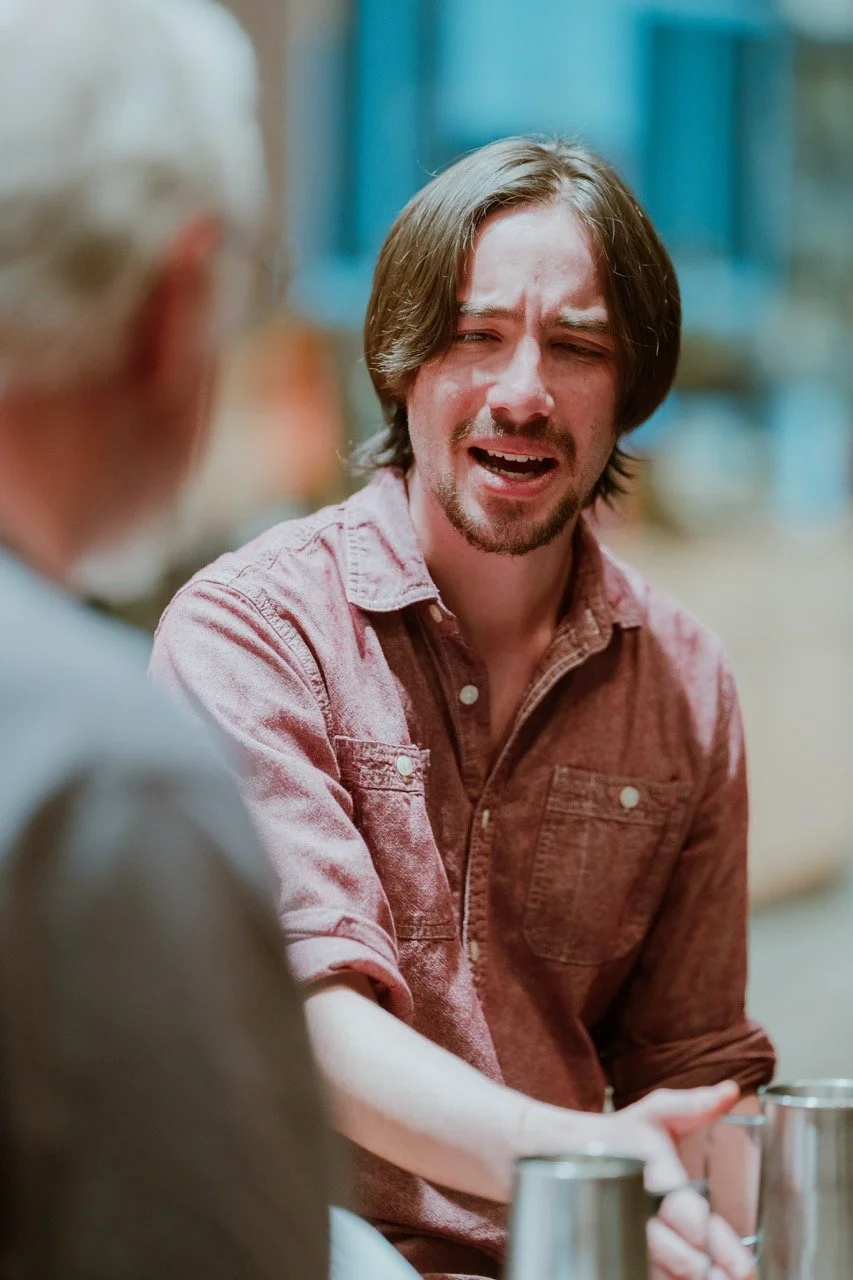 A man with long hair and a beard wearing a red shirt is sitting and gesturing, appearing to be in conversation. Another person is out of focus in the foreground.