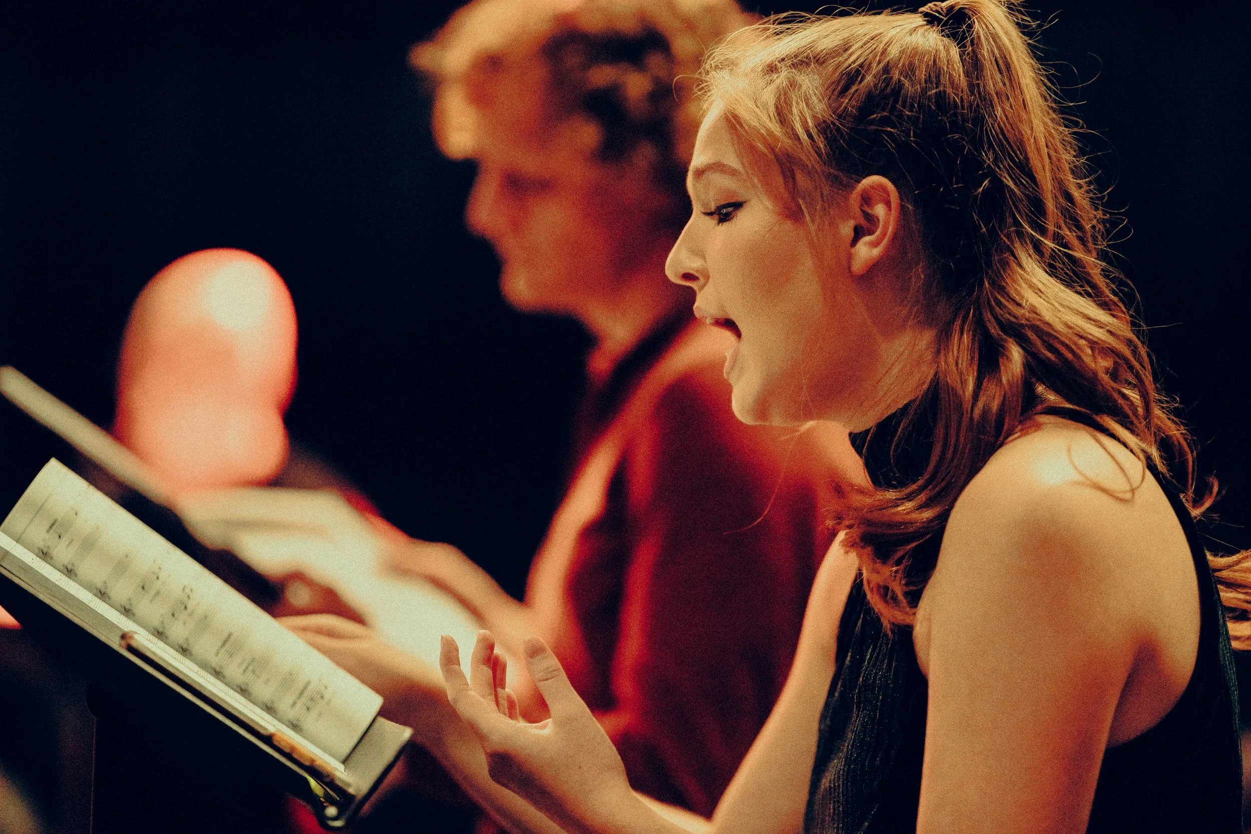 Woman singing with a choir during a rehearsal, holding sheet music, with another person in the background.