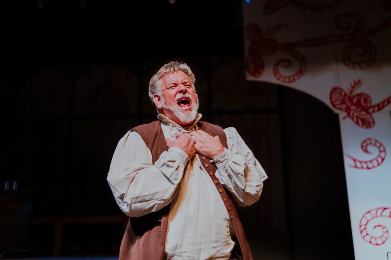 An actor performing on stage, wearing a period costume with a vest and shirt, expressing an emotional moment.