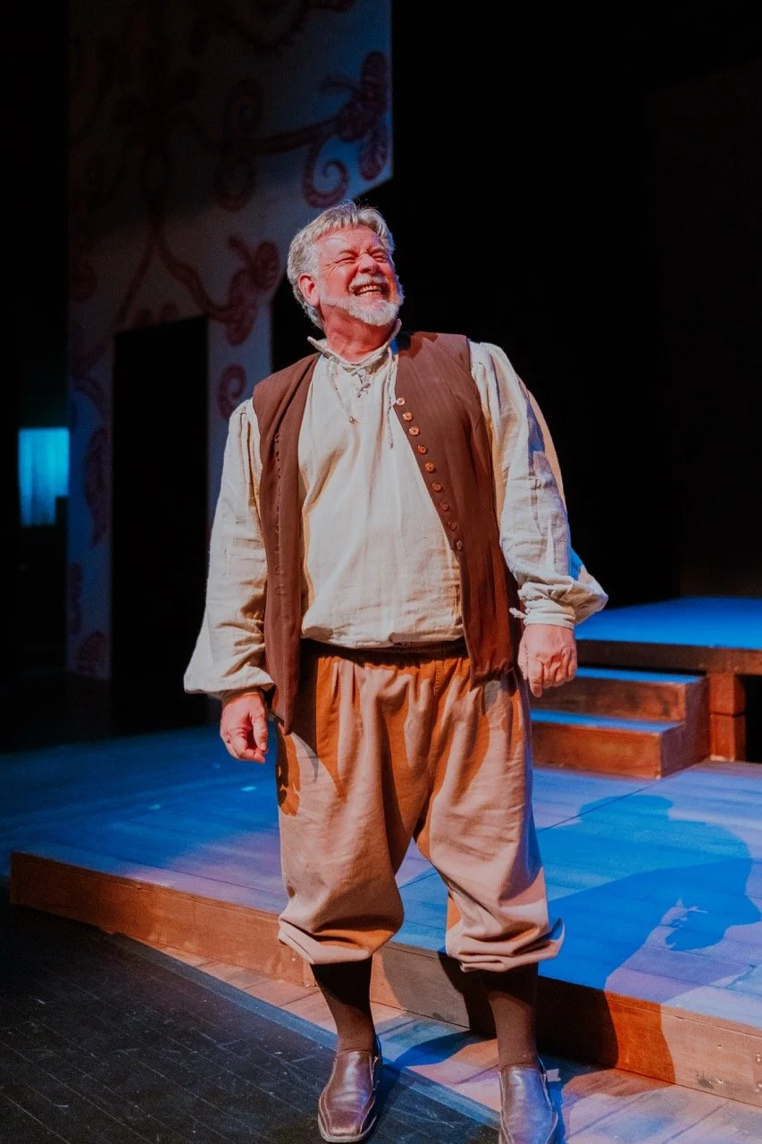 Man in historical costume smiling on stage, wearing a white shirt, brown vest, and beige pants.