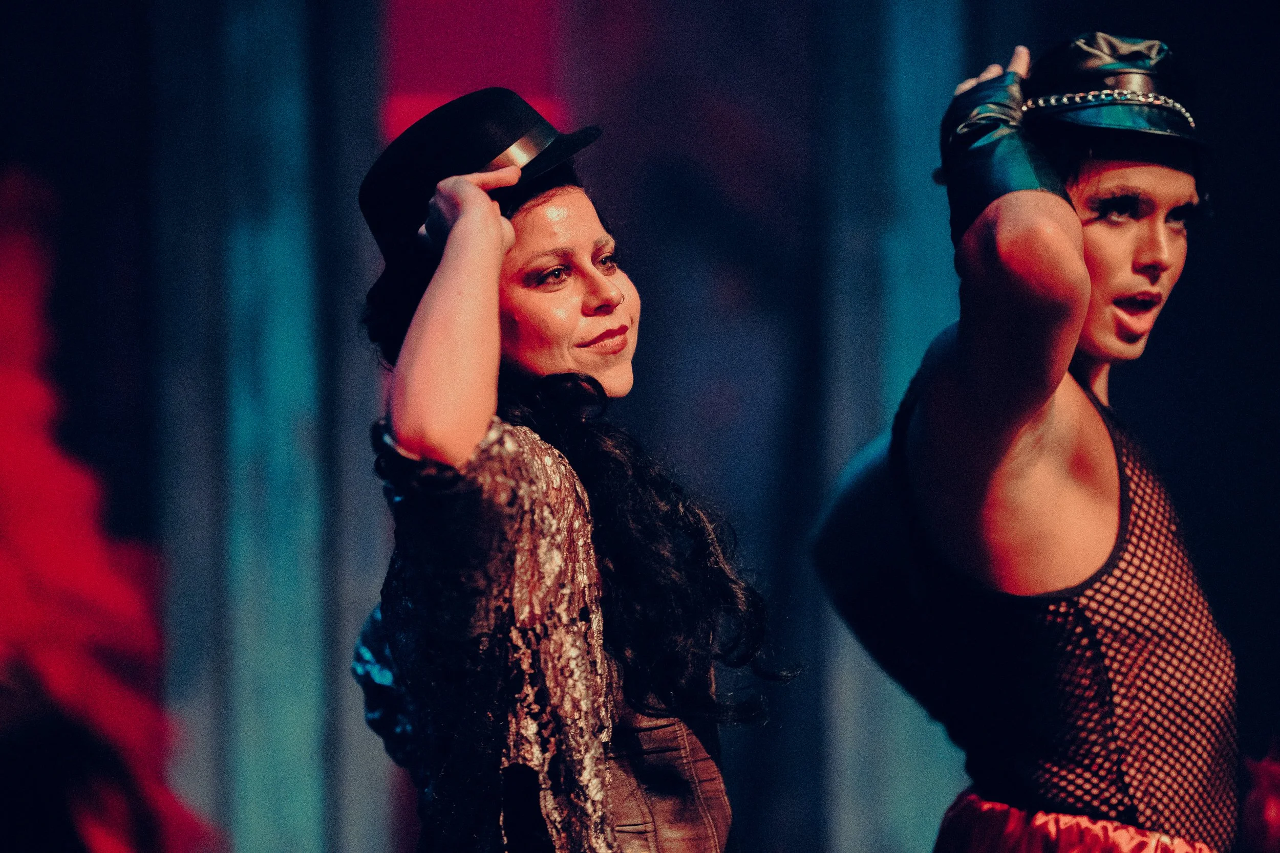Two performers on stage wearing costumes and hats, one in a sequined top and the other in a mesh outfit, with dramatic lighting.
