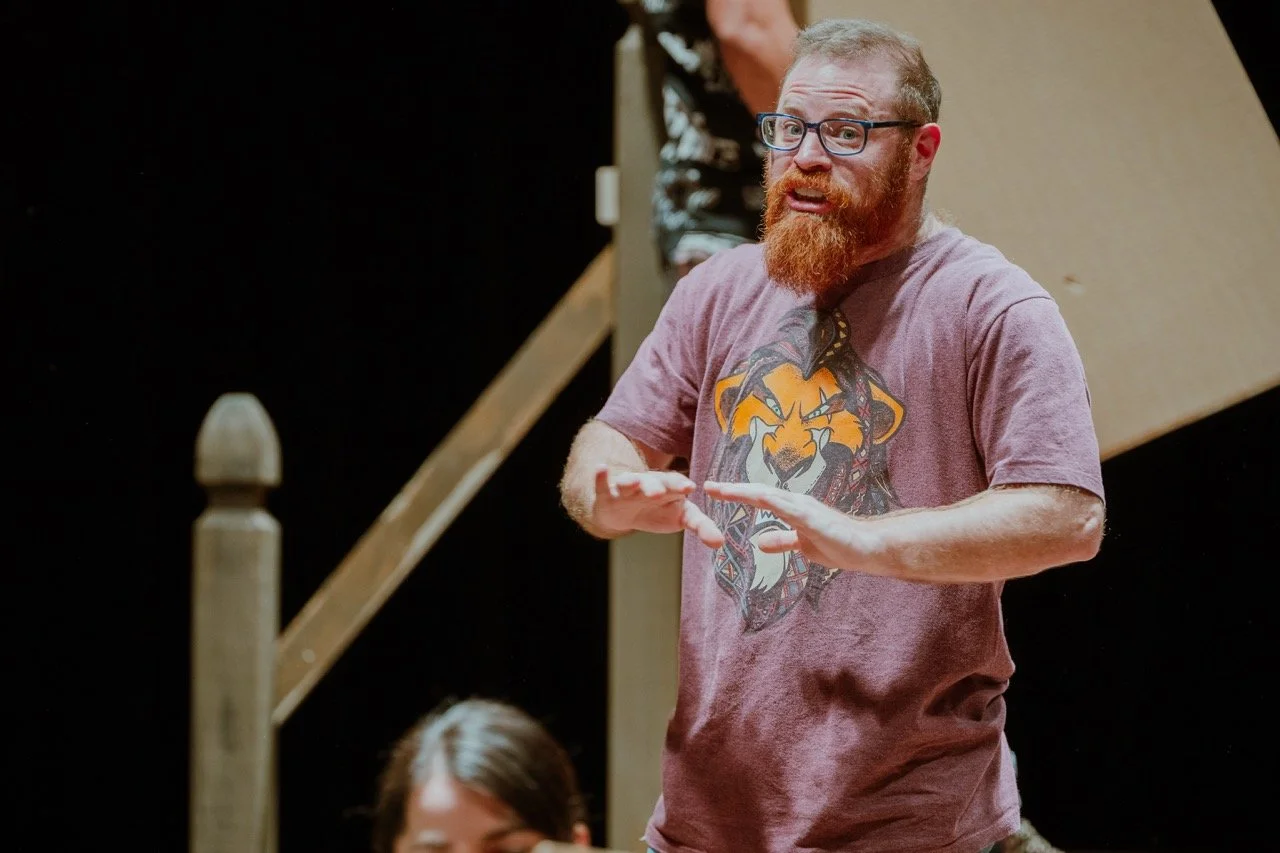 A person with a beard and glasses wearing a T-shirt featuring a lion design, speaking or gesturing on stage.