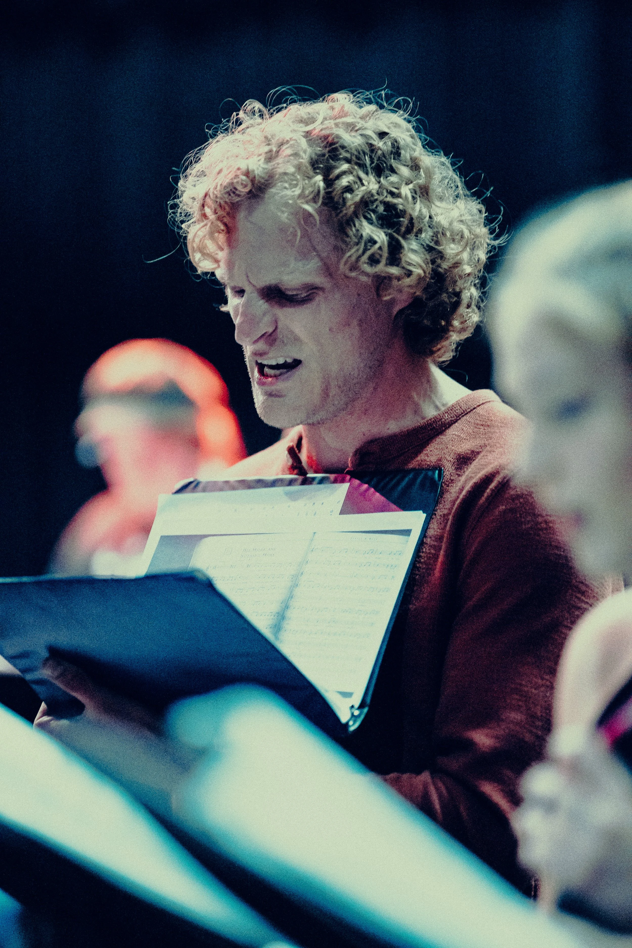 Person on stage holding and reading from sheet music, appearing focused and engaged.