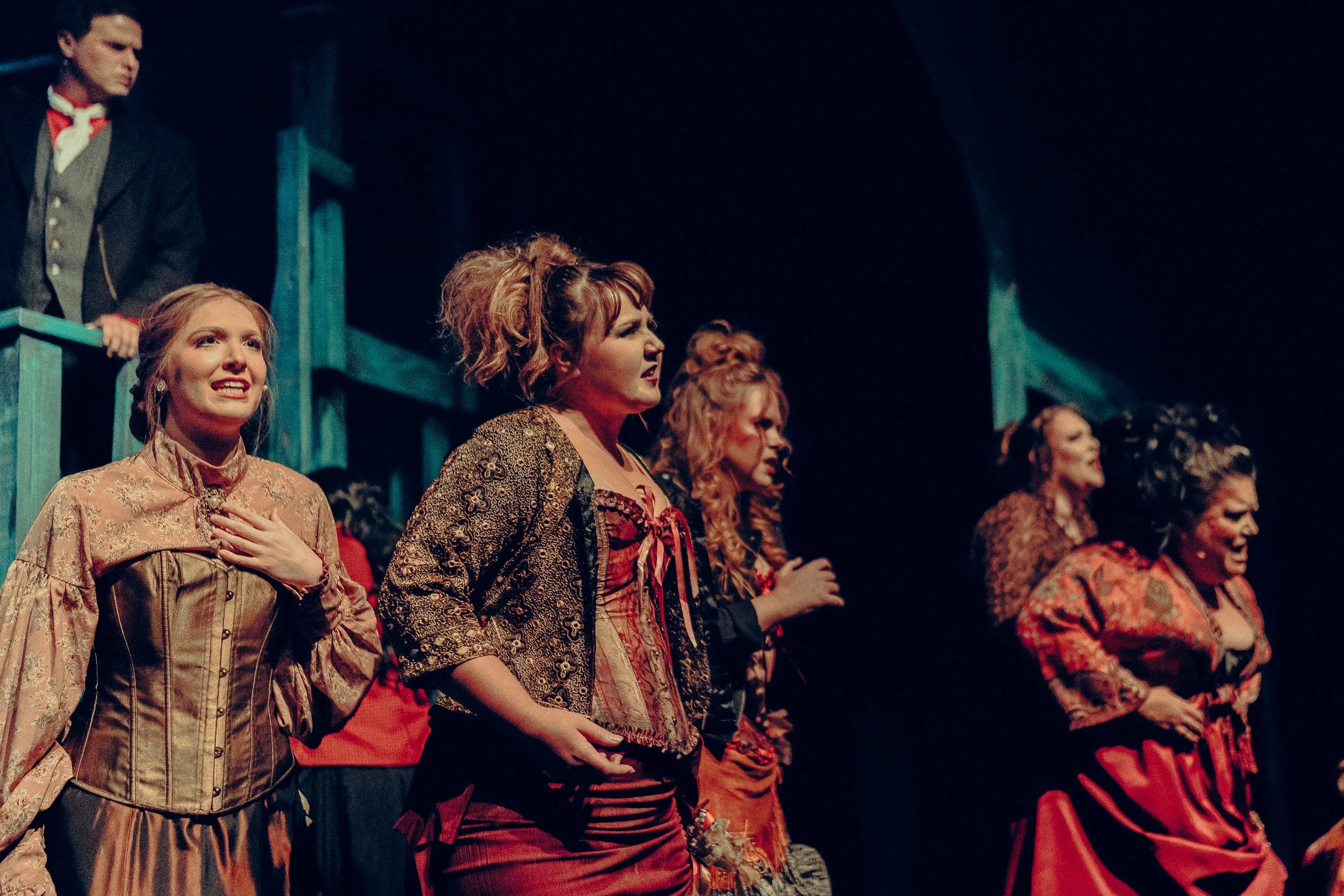 Theater performers on stage in period costumes, including women wearing elaborate dresses and corsets, during a dramatic musical scene with dim lighting and a dark backdrop.