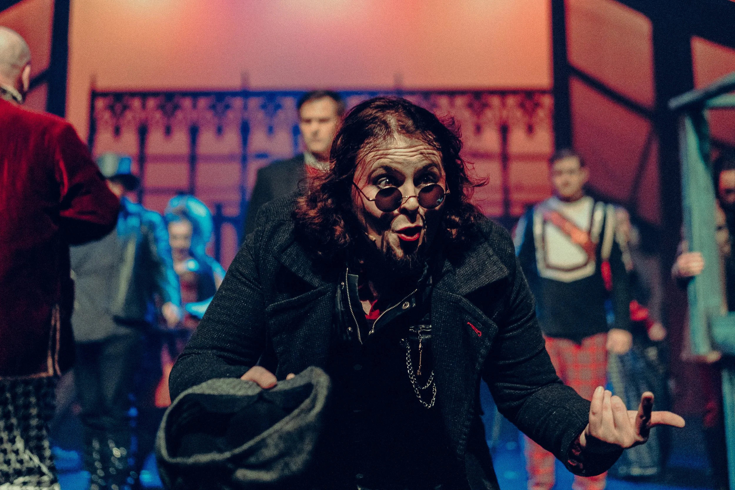 Stage performance with a person in dark clothing and sunglasses, energetically gesturing with others in costume behind.