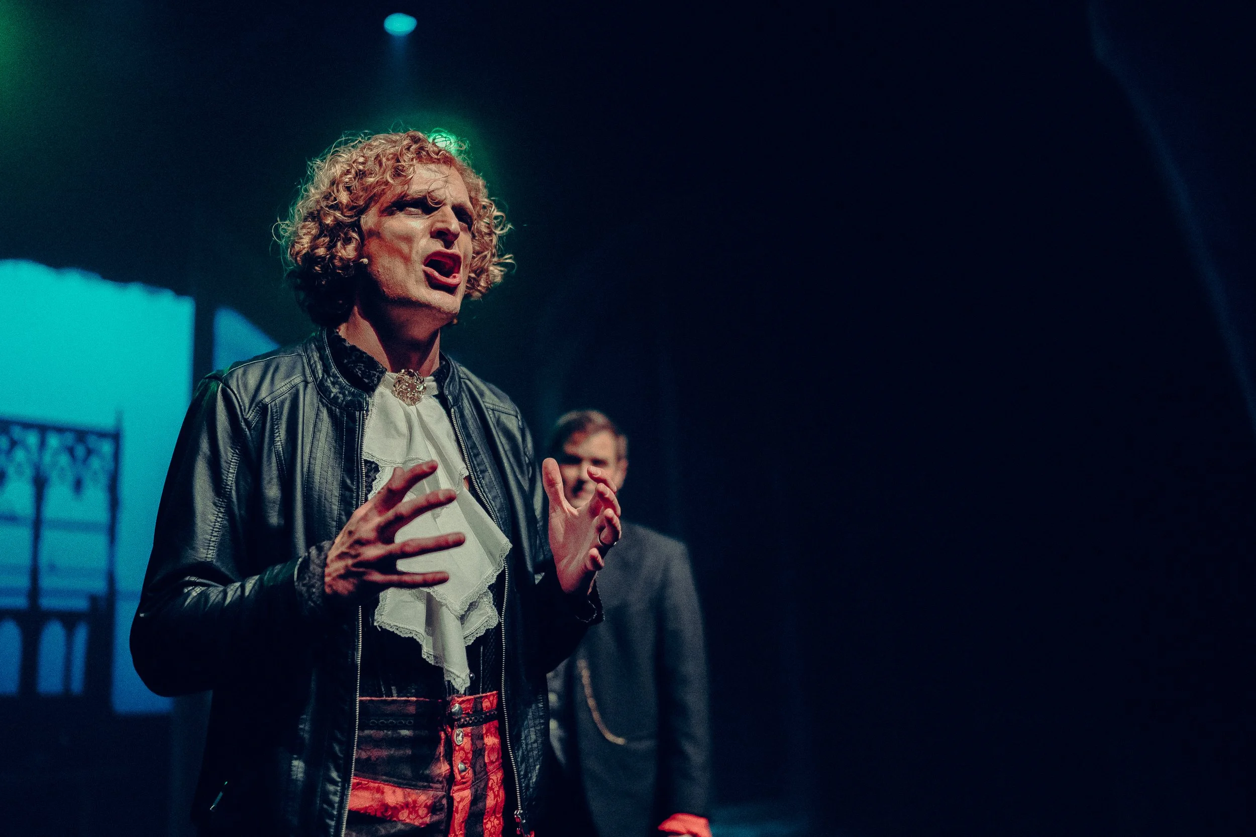 Man with curly hair in theatrical performance, wearing a black jacket and white ruffled shirt, expressive gestures, with stage lighting and another performer in the background.