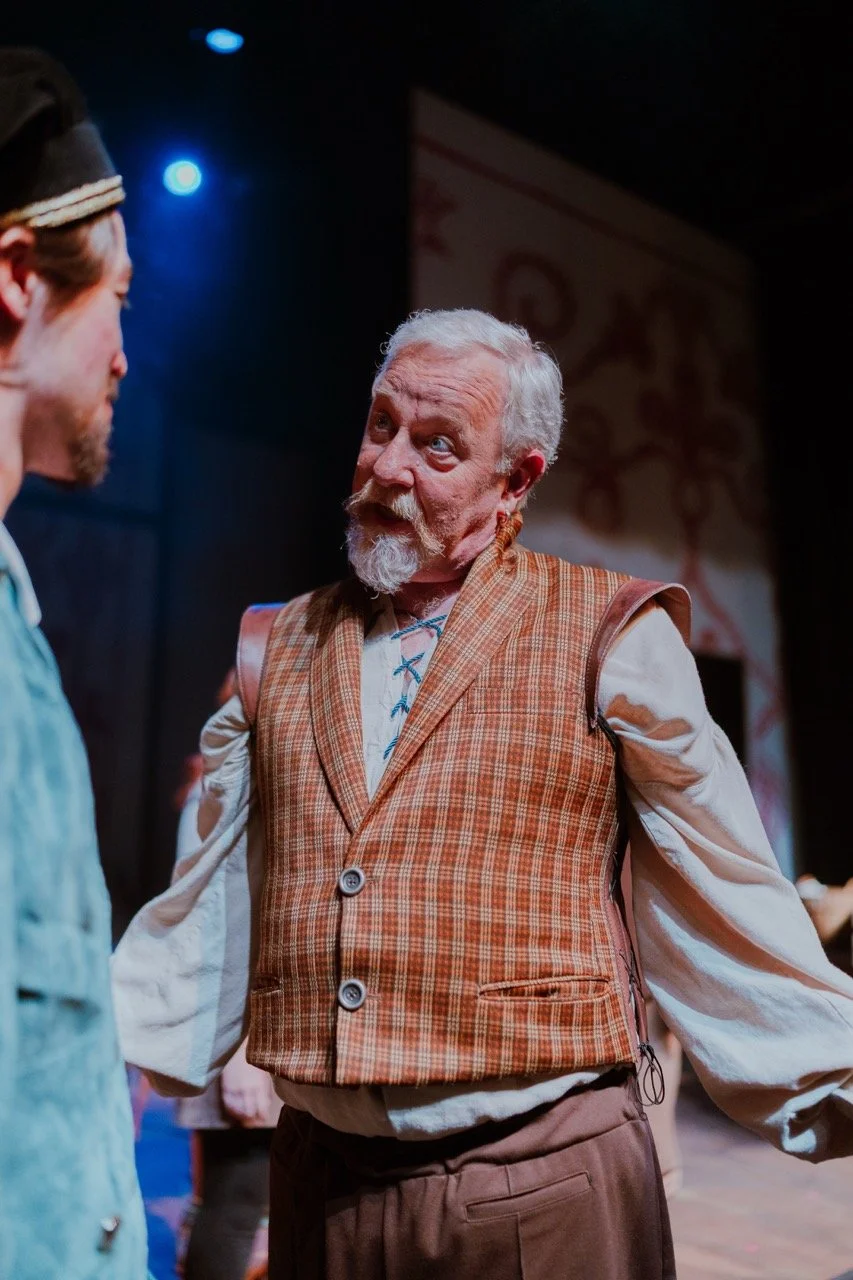 An older man with a white beard and hair wearing a plaid vest and a white shirt, performing on stage during a play, with theatrical lighting highlighting him.