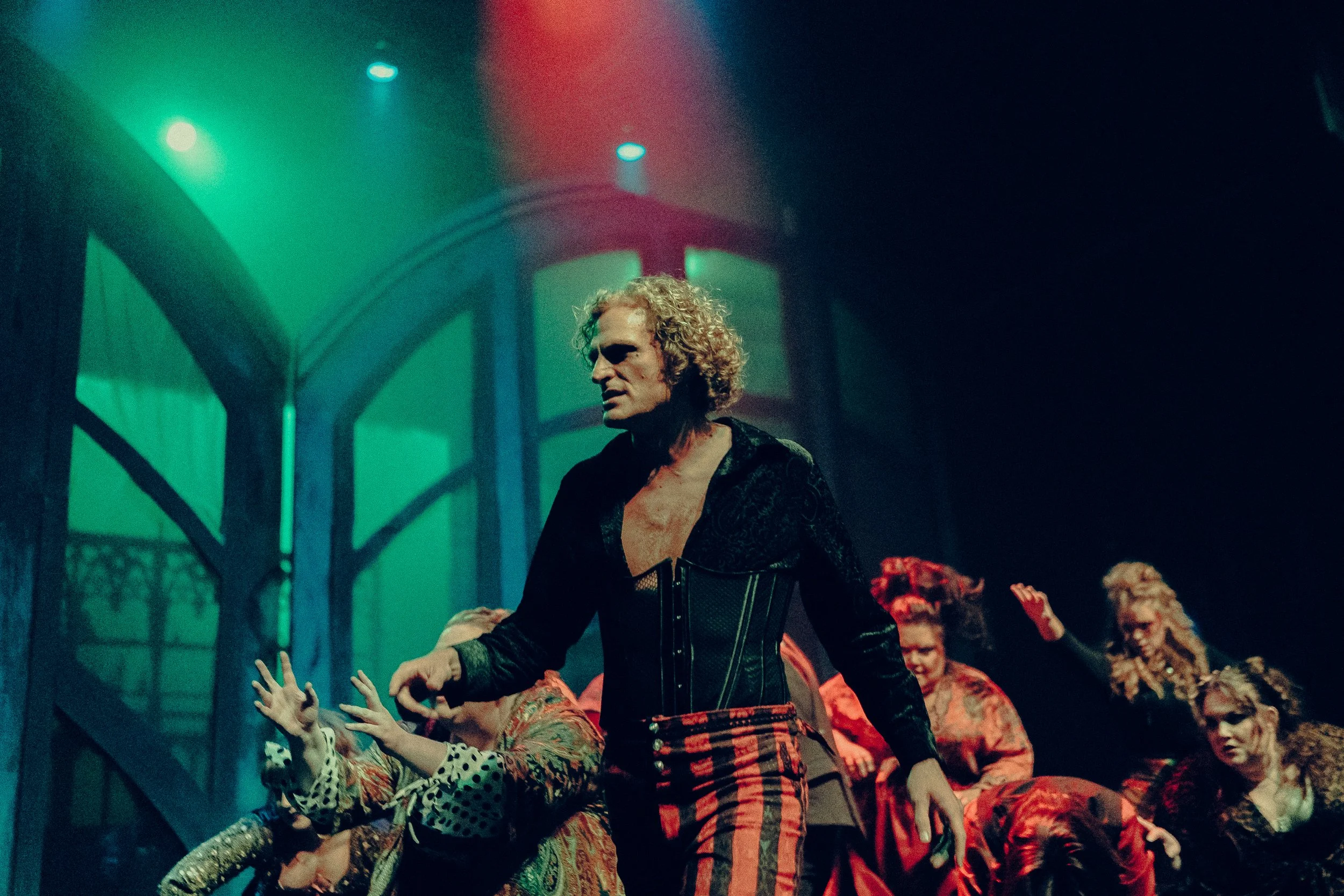 A theatrical performance with dramatic lighting, featuring a man in an elaborate costume of a black top and red-striped pants, with performers in dynamic poses in the background.