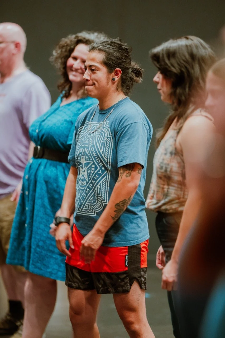 Group of people standing in a line, listening attentively in casual clothing, with focus on a person in a blue t-shirt and red shorts.