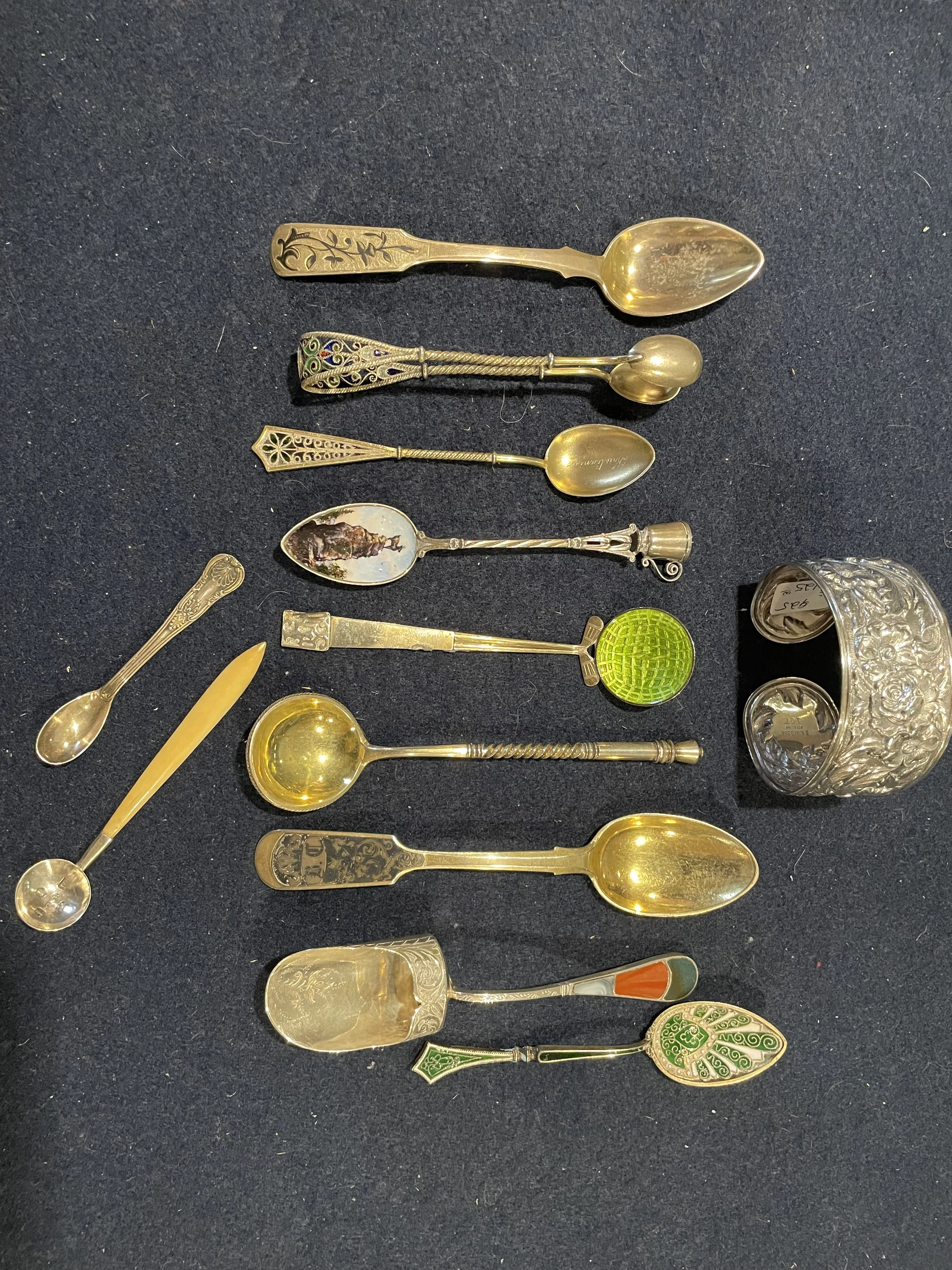 A rare assortment of gold-washed and sterling collector spoons, featuring vibrant plique-à-jour enamel, hand-painted scenics, and a stunning heavy floral repoussé cuff bracelet