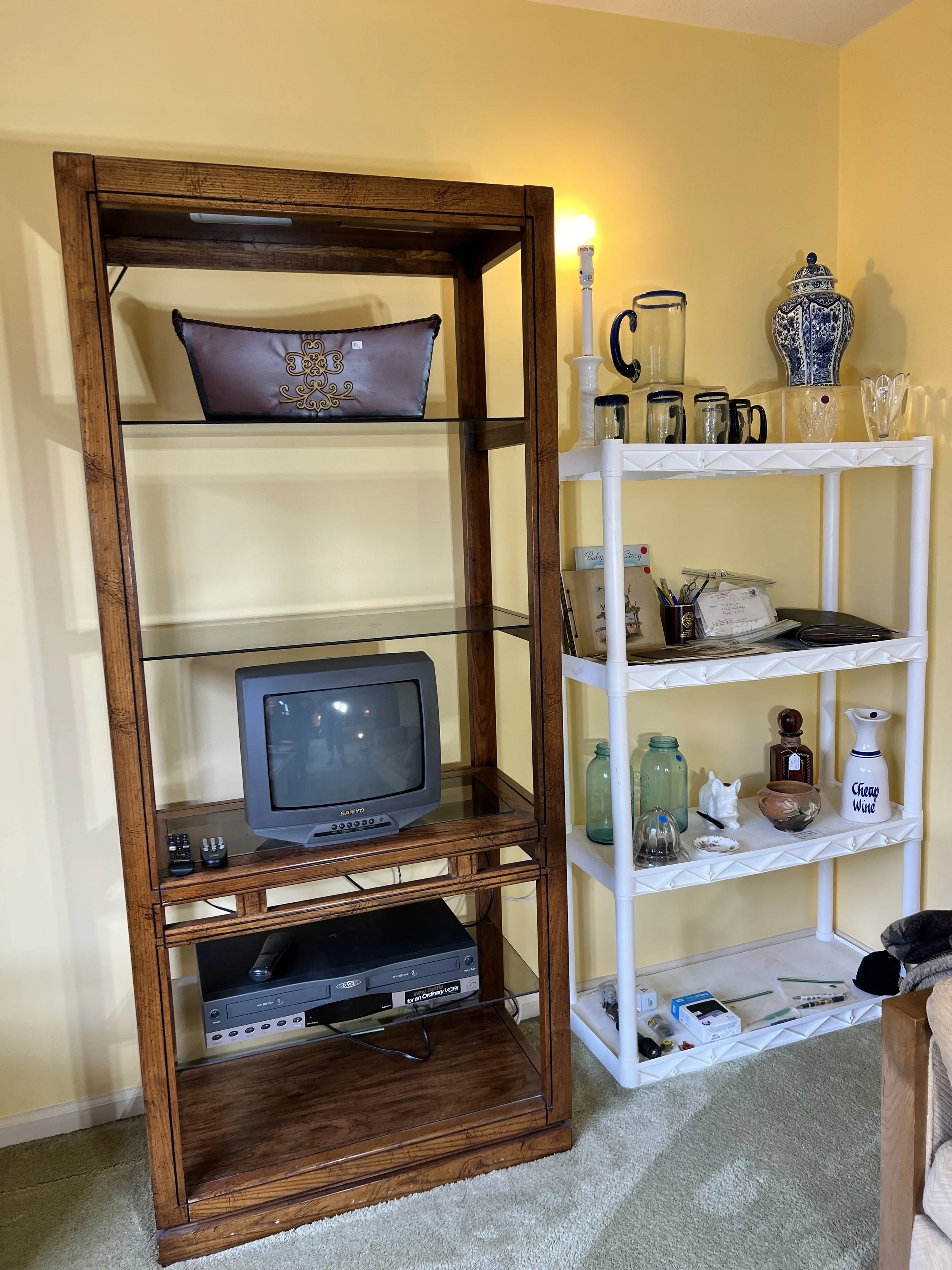 A tall wood-frame glass etagere housing a Sanyo CRT TV and VCR, barware, and antique glass jars