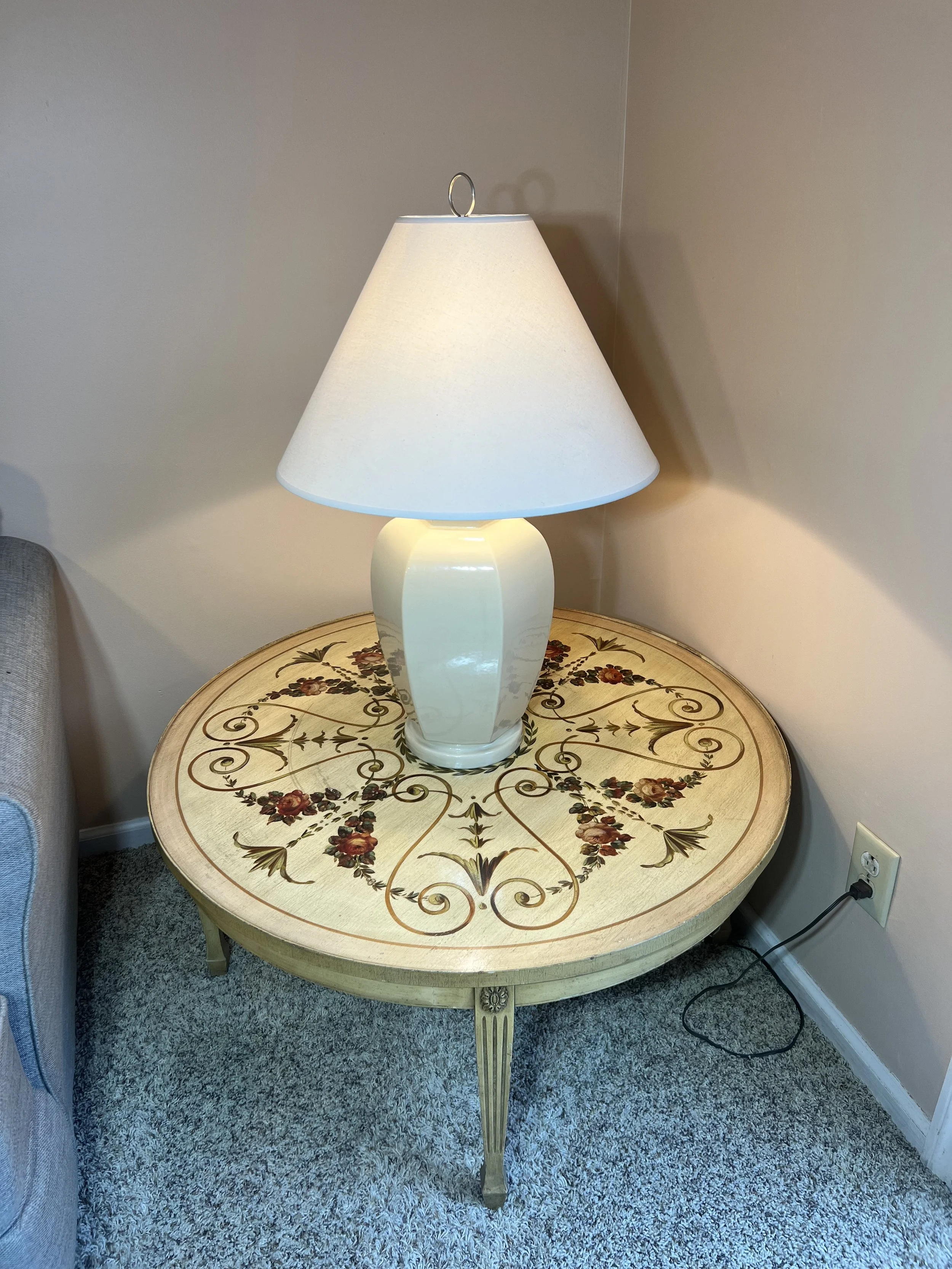 A round, cream-painted end table featuring an intricate hand-painted floral and scrollwork design and lamp with shade