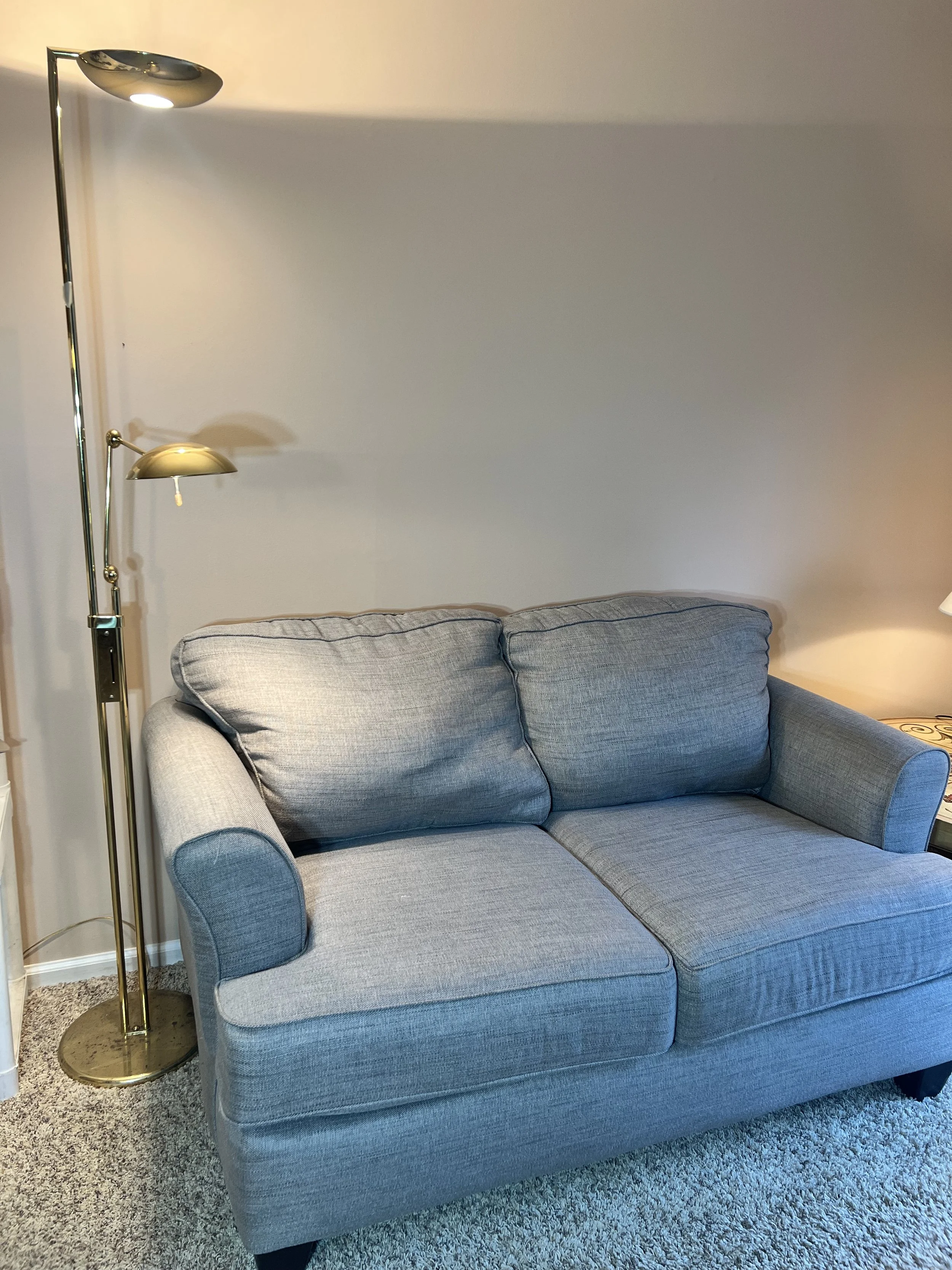 A tall, sleek brass floor lamp with an adjustable "pharmacy style" arm, shown next to a plush gray loveseat