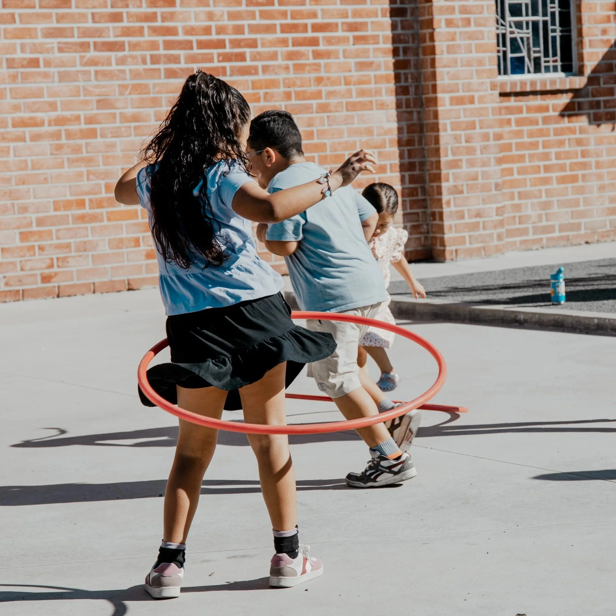 Three children playing with a hula hoop outdoors on a sunny day, standing against a red brick wall.