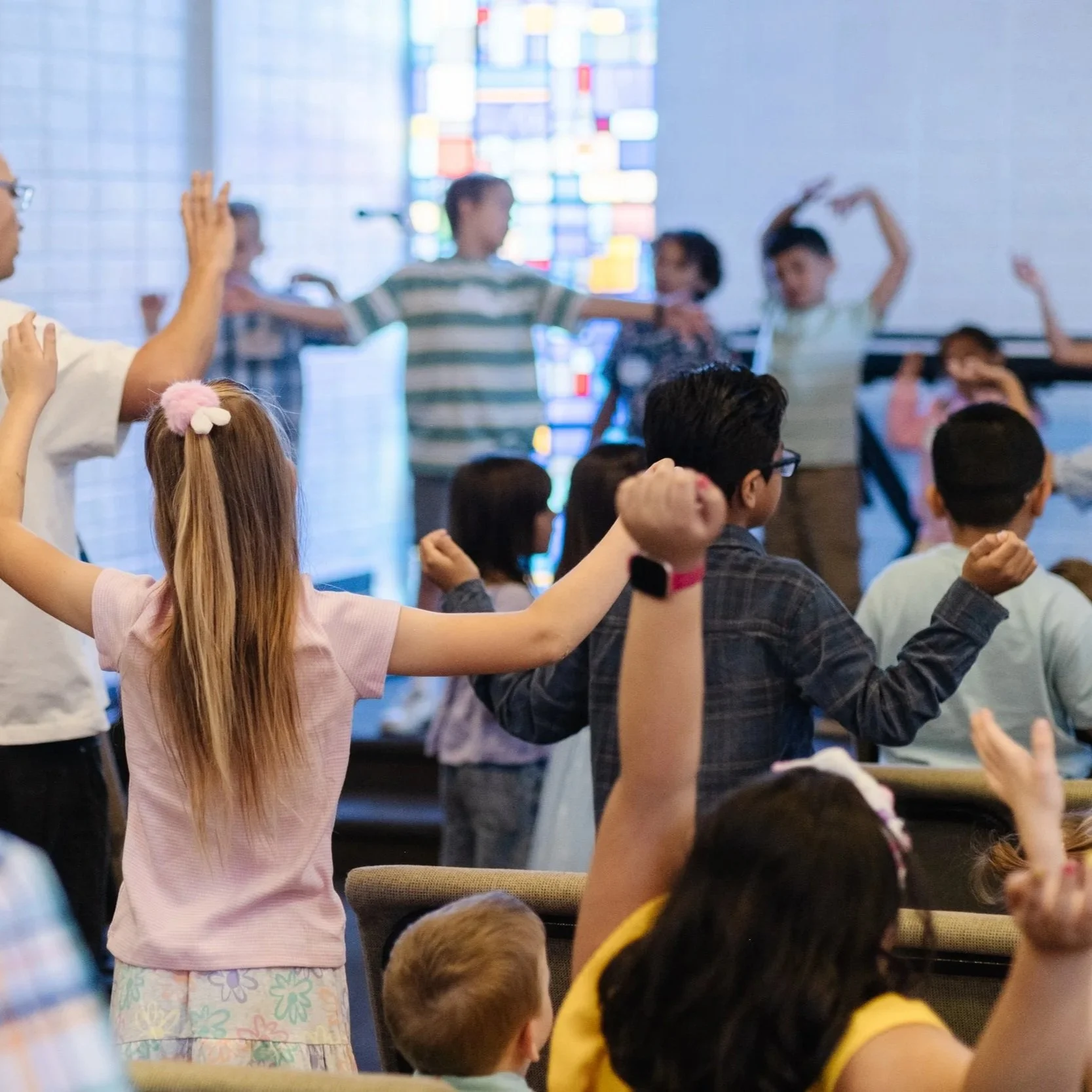 A group of children and adults participating in a dance or exercise class in a well-lit room with a colorful abstract window in the background.