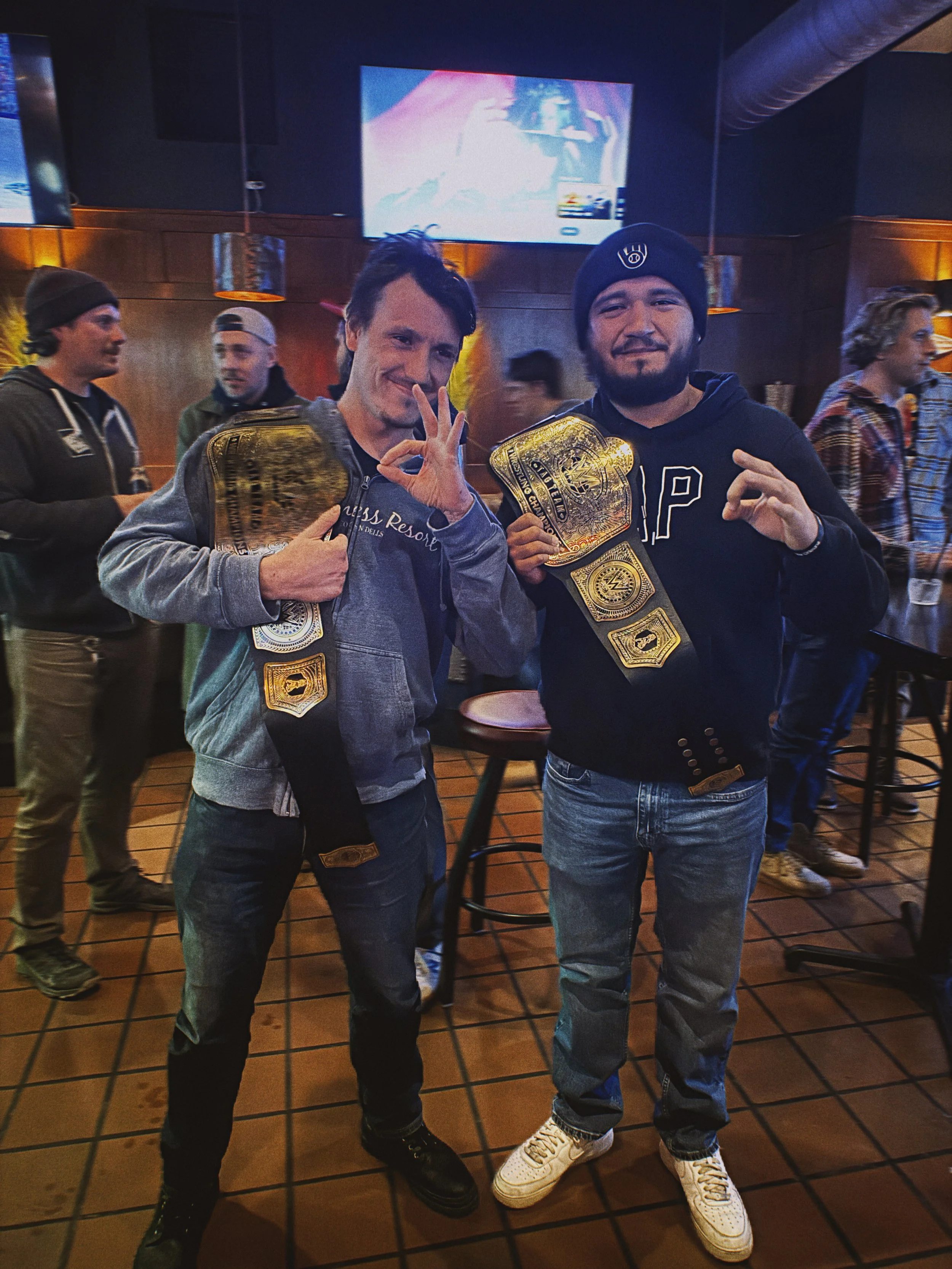 Two men standing in a bar Pete's Pub holding WWE replica championship belts won in a giveaway, smiling, with several people in the background.