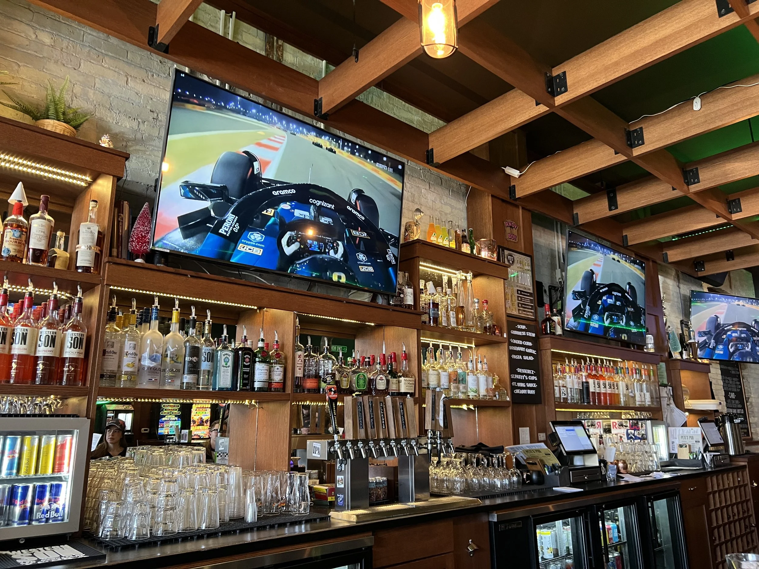 Multiple flat screen TVs showing a formula one F1 Grand Prix race live with sound in a bar in Milwaukee, Wisconsin with wooden shelves stocked with bottles of alcohol and glasses, and a brick wall background.