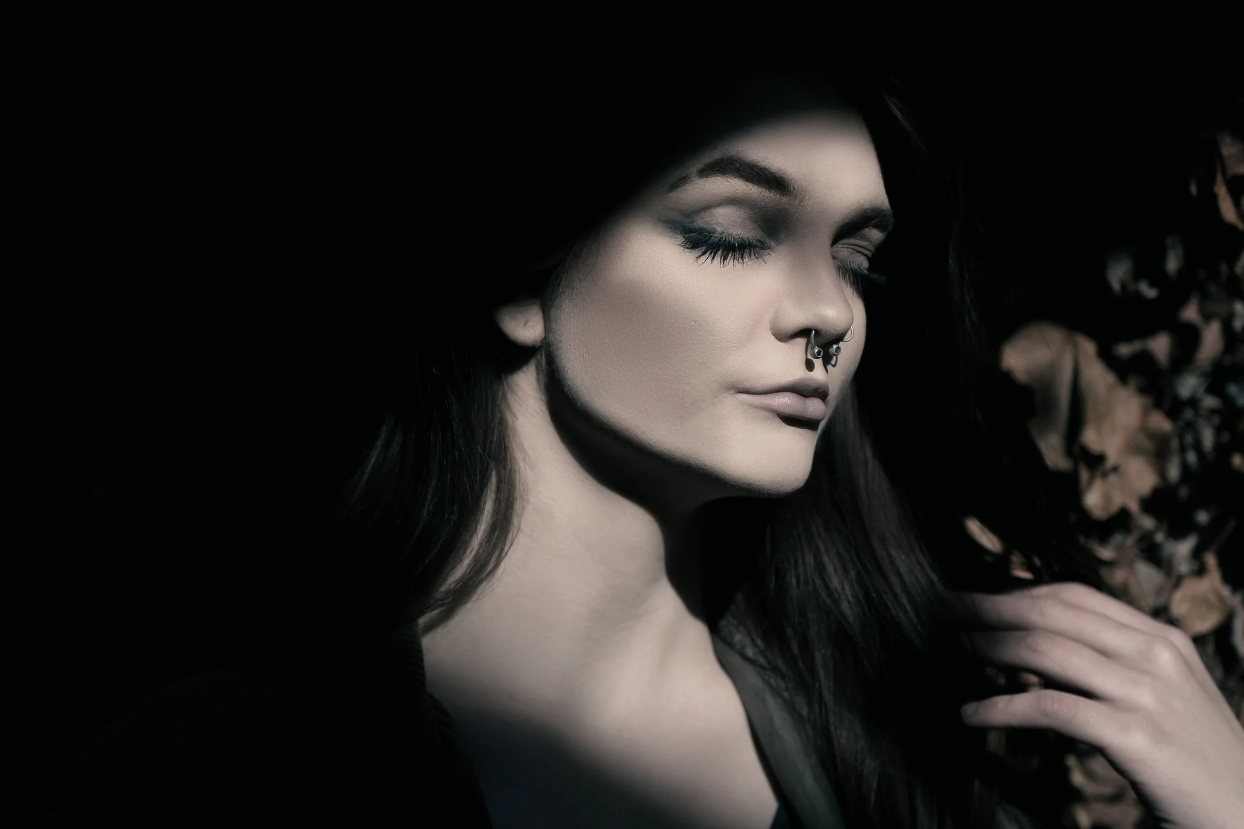A young woman with black hair, closed eyes, septum piercing, and makeup, resting her hand on her shoulder in a dimly lit environment.