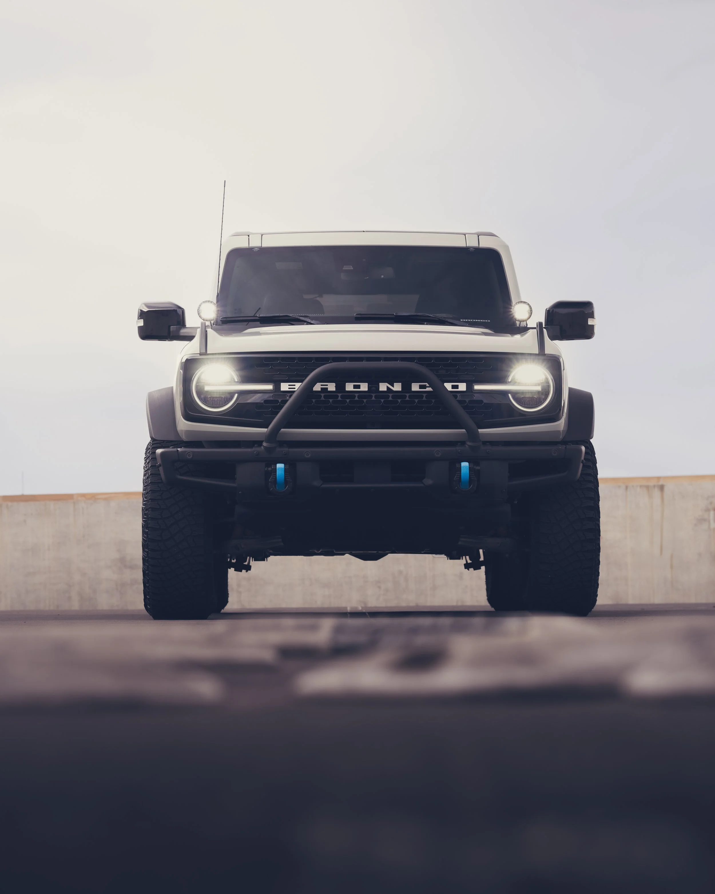 Front view of a black Ford Bronco SUV with illuminated headlights, parked on a rooftop against a cloudy sky.