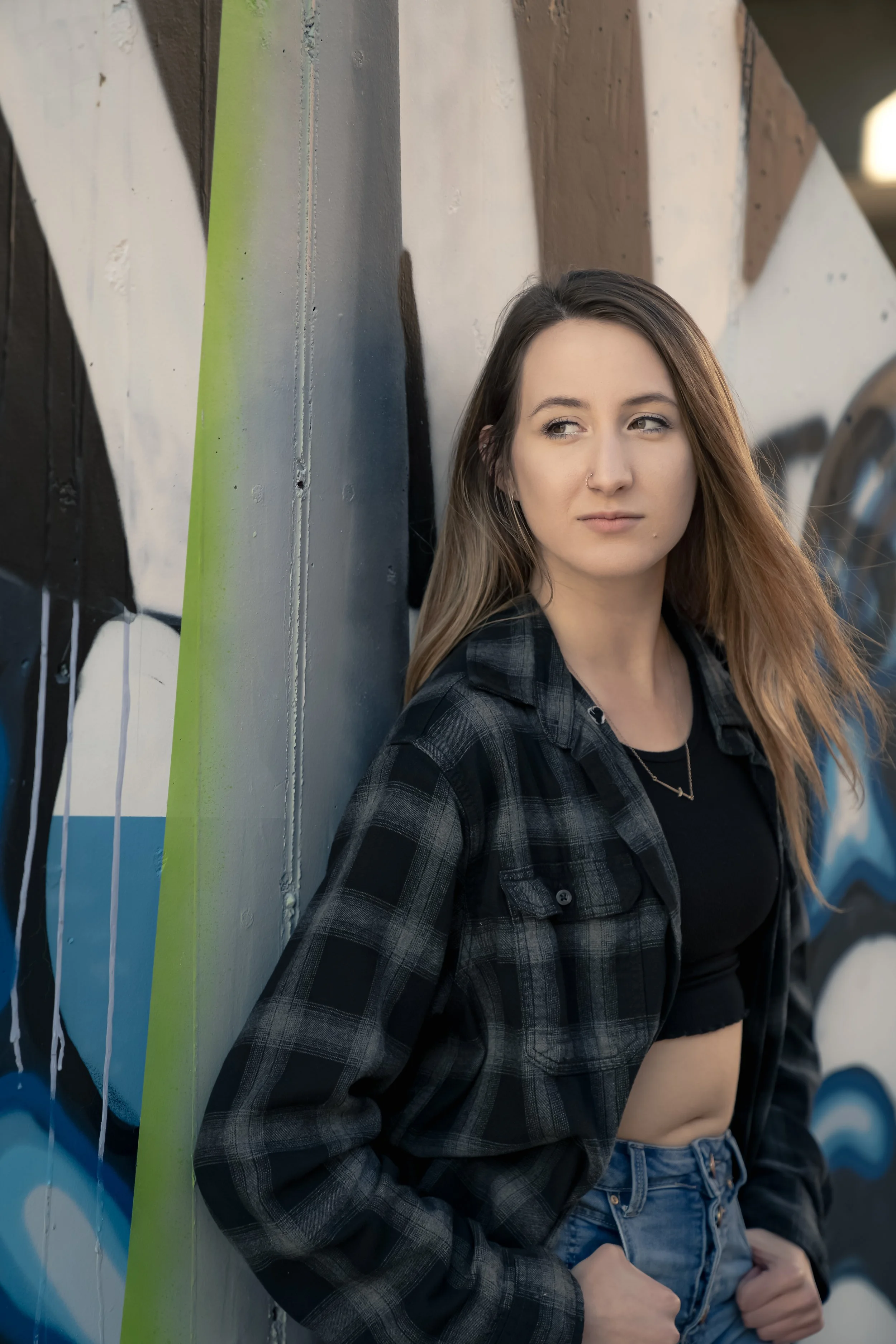 A young woman with long light brown hair wearing a black crop top and black plaid shirt stands against a graffiti wall.