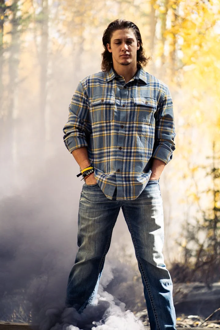 A young man with long hair and a serious expression, standing outdoors amidst smoke or fog, wearing a blue plaid shirt and jeans, with autumn trees in the background.
