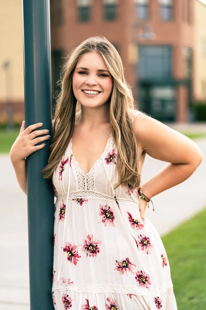 Young woman with long blonde hair smiling, leaning on a blue pole outdoors in a casual summer dress with pink floral pattern, in front of a city building.