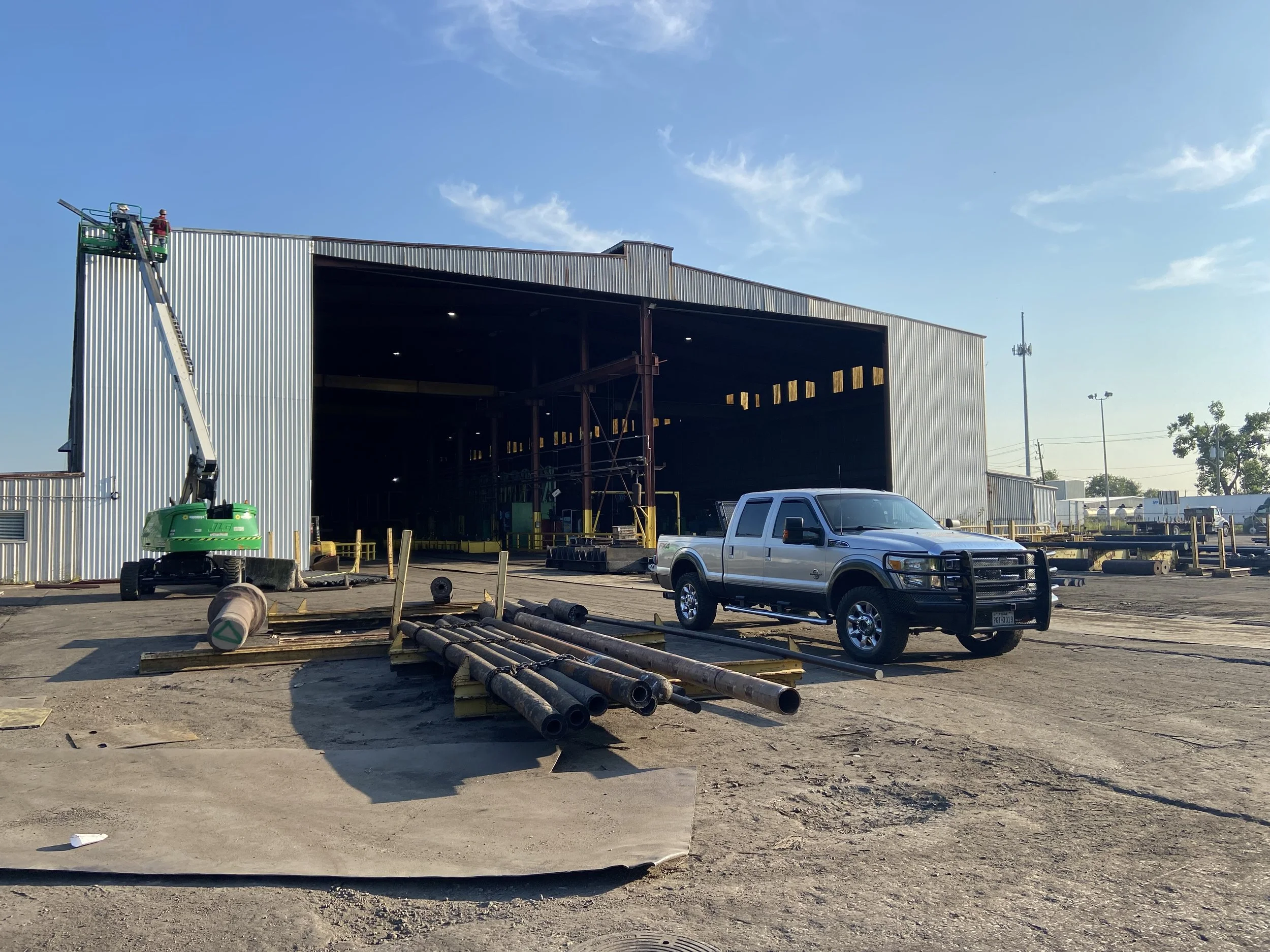Construction site with a large partially built metal building, a silver pickup truck parked in front, and construction pipes and equipment on the ground under a clear blue sky.