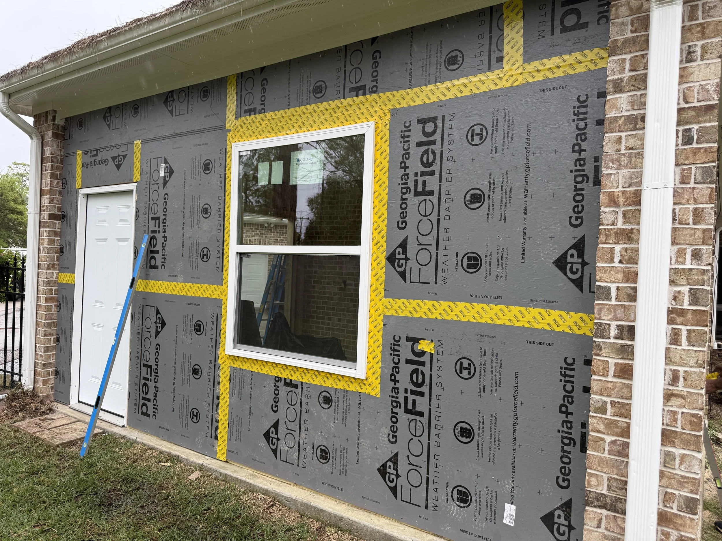 Exterior of a house under construction with siding being installed. The house has brick walls, a white window, a white door, and a white rain gutter. Insulation and weather-resistant barrier wrap are visible on the exterior wall.