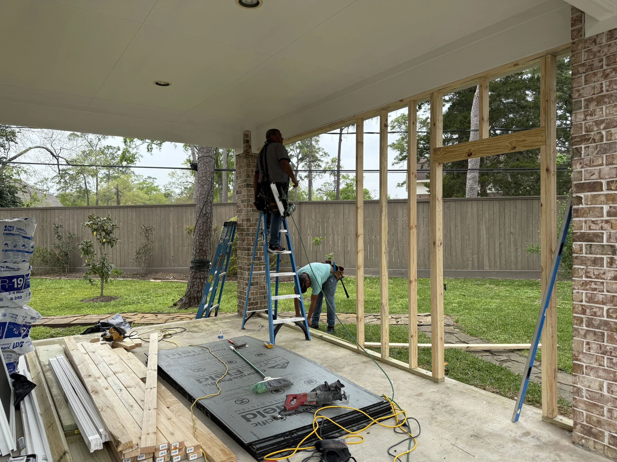 Two construction workers building a wall on a covered porch, with exposed wooden framing, tools, and construction materials on the floor.