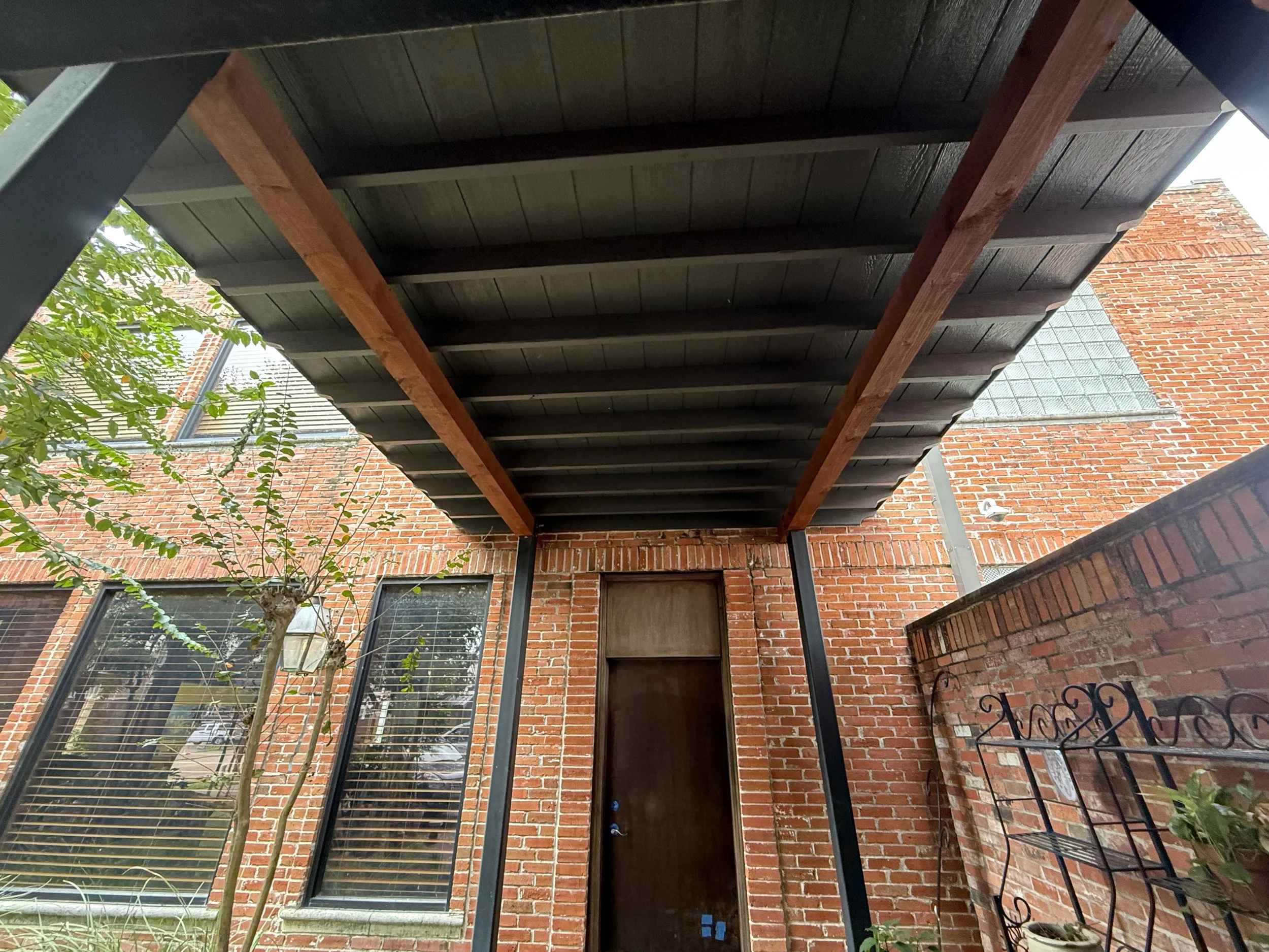 View of a brick building with two large windows, a wooden door, and a black metal railing and plant on a small patio. The patio has a covered roof with black panels and wooden beams.