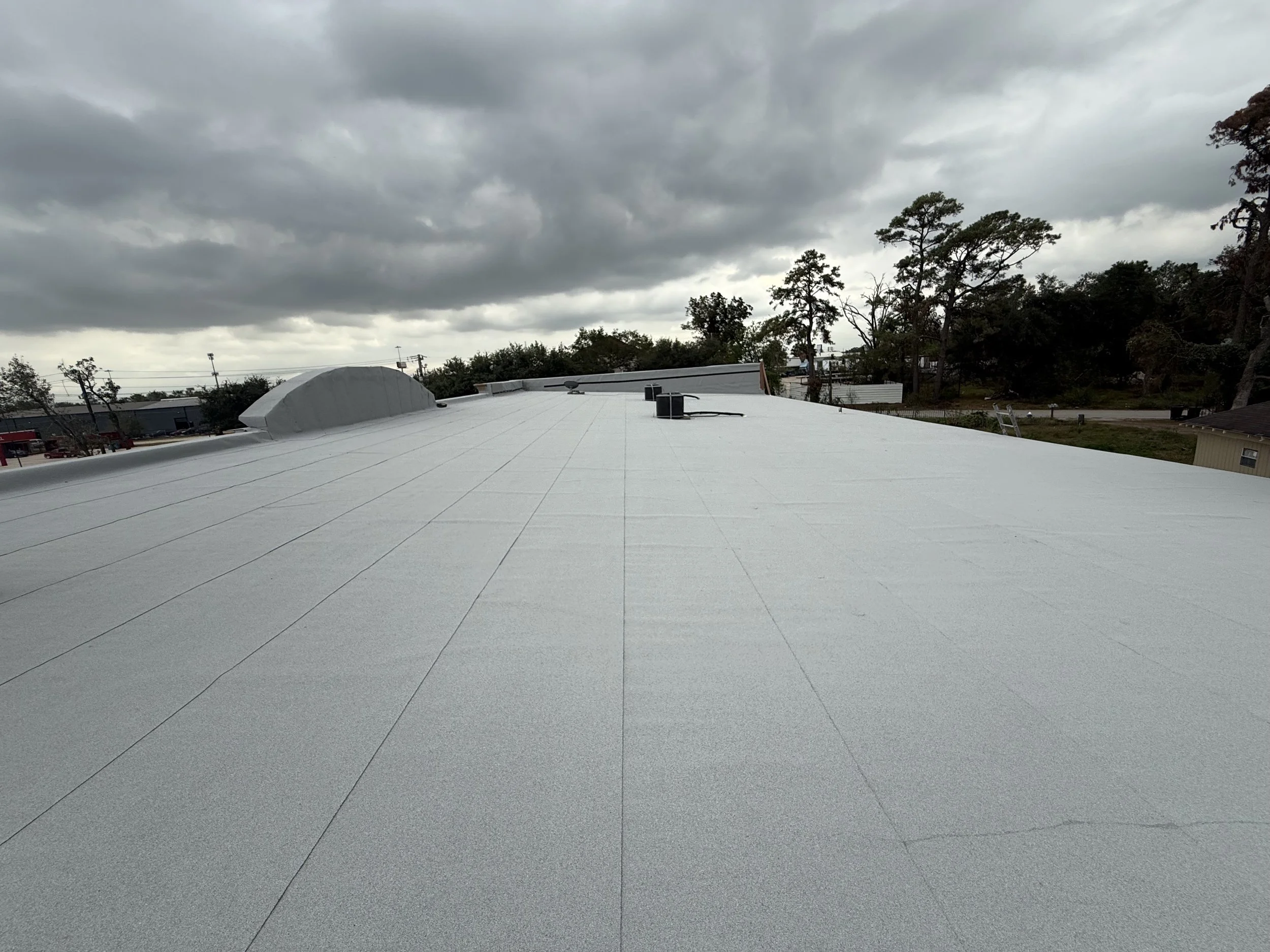 A flat white commercial roof with asphalt shingles, some vents and a small curved skylight, under a cloudy sky with trees and buildings in the background.