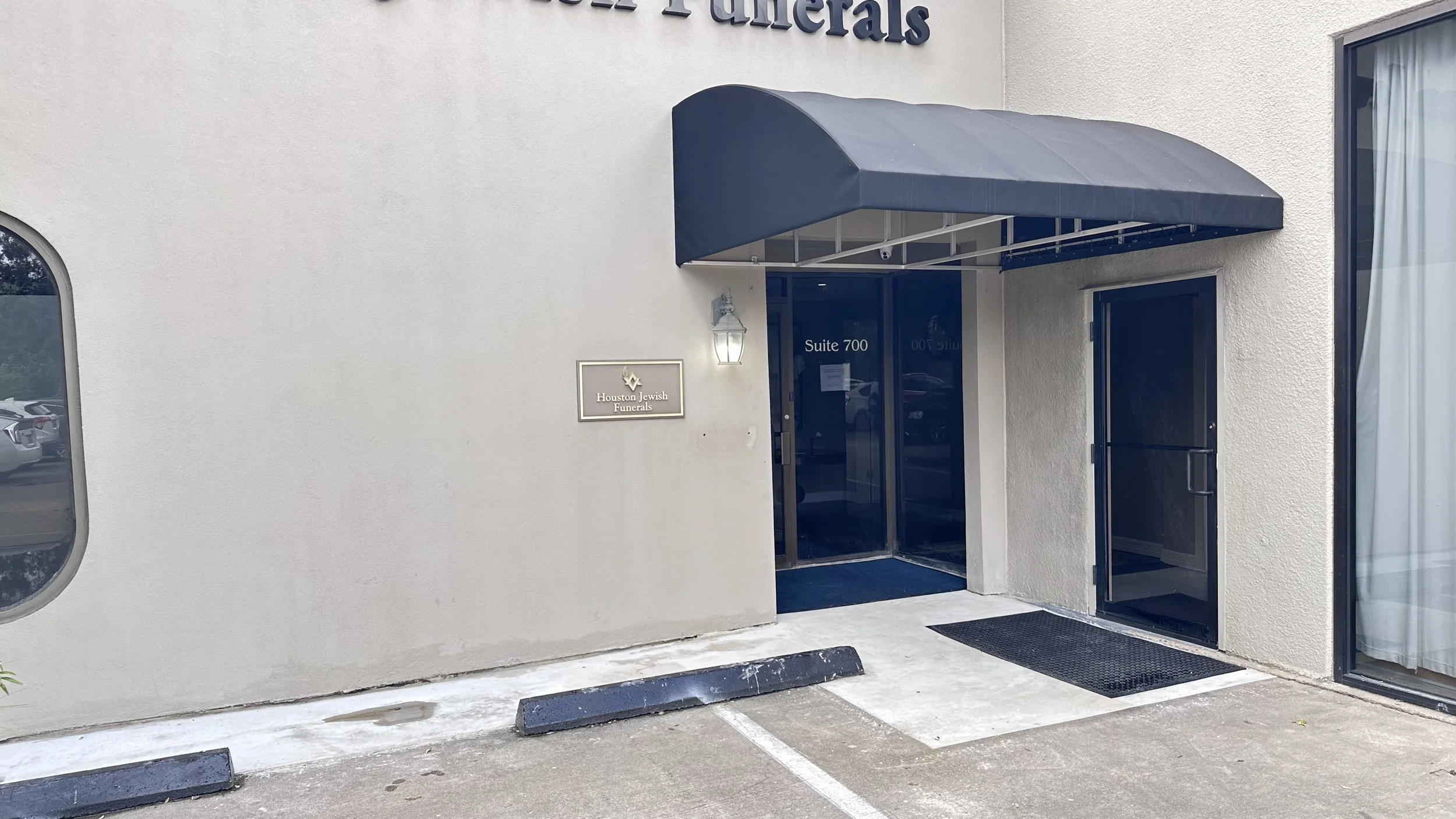 Exterior view of Houston Jewish Funerals building with a black awning over the entrance, a wall-mounted lantern light, and a sign indicating the location and suite number.