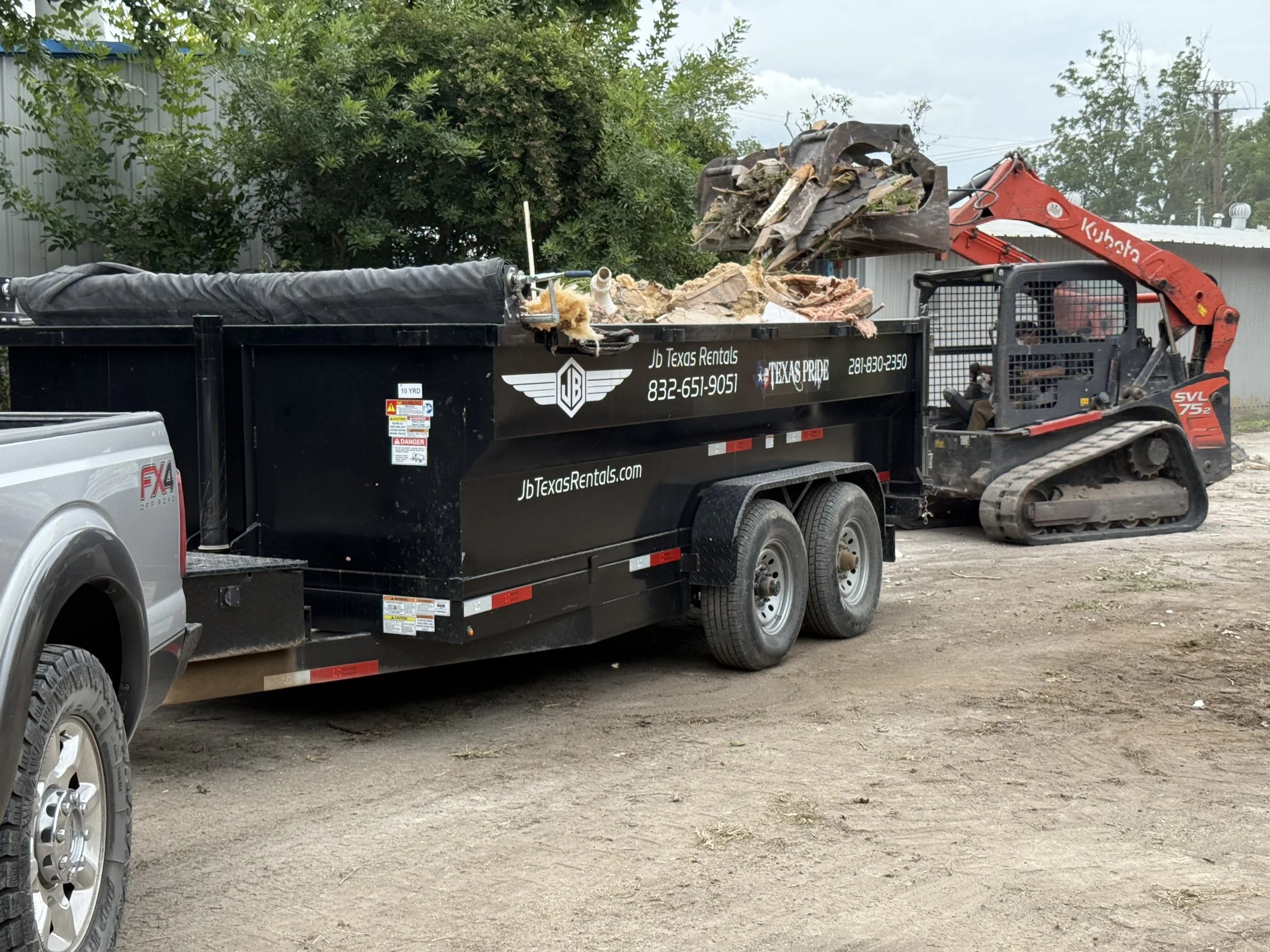 A black trailer attached to a pickup truck, loaded with debris, with a small red excavator on dirt ground in a yard.