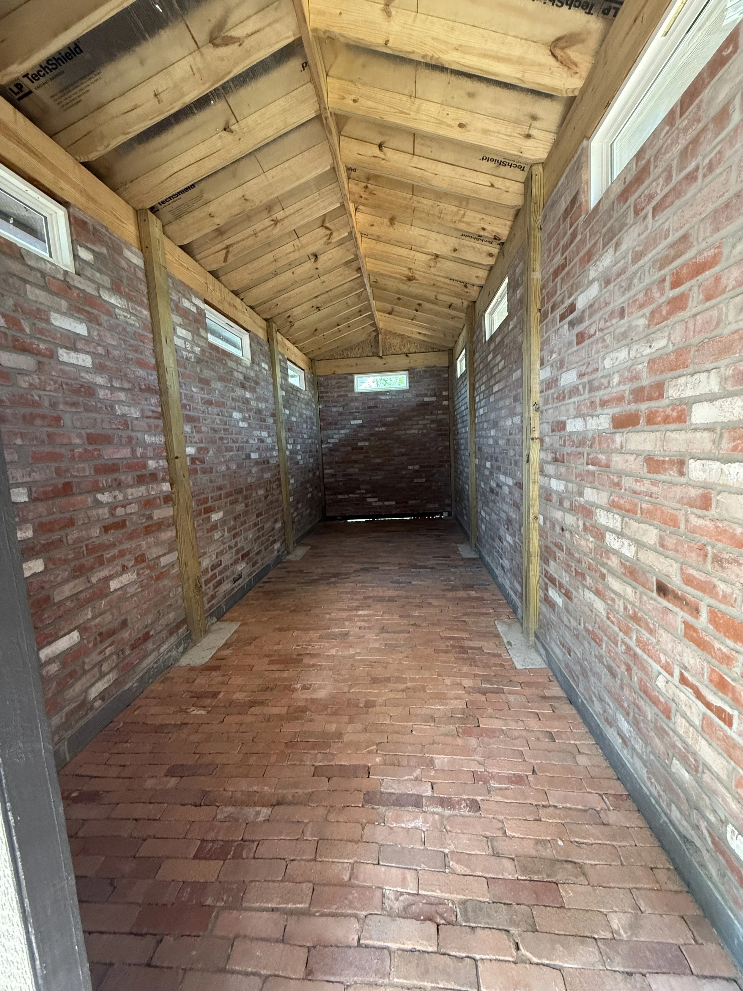 A covered outdoor walkway with brick flooring, brick walls, small rectangular windows, and a wooden ceiling with visible insulation.
