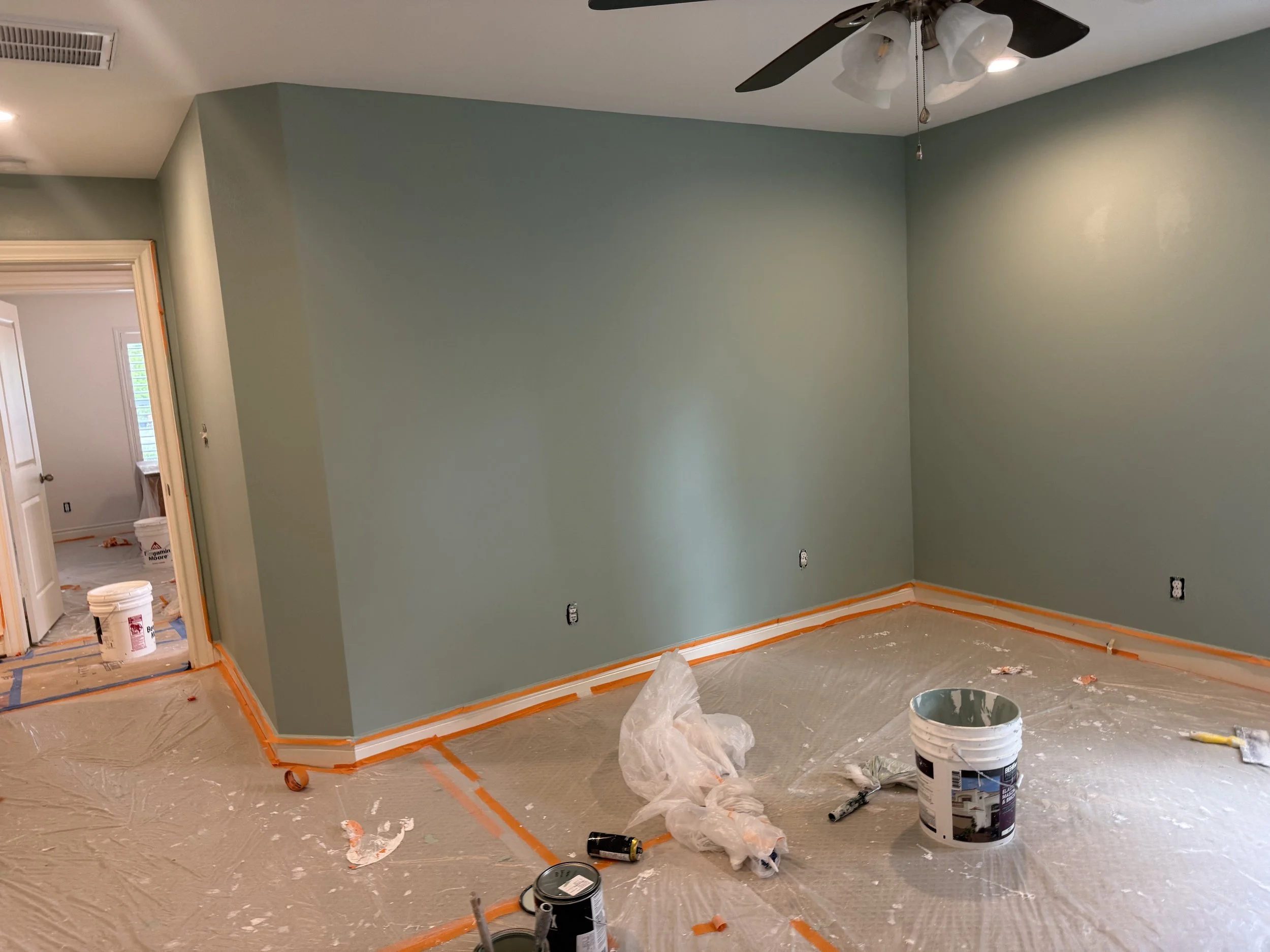 Room under renovation with green painted walls, plastic covering the beige carpeted floor, and orange painter's tape outlining the floor edges. There are paint supplies, including buckets and brushes, scattered around. A ceiling fan is visible, and a