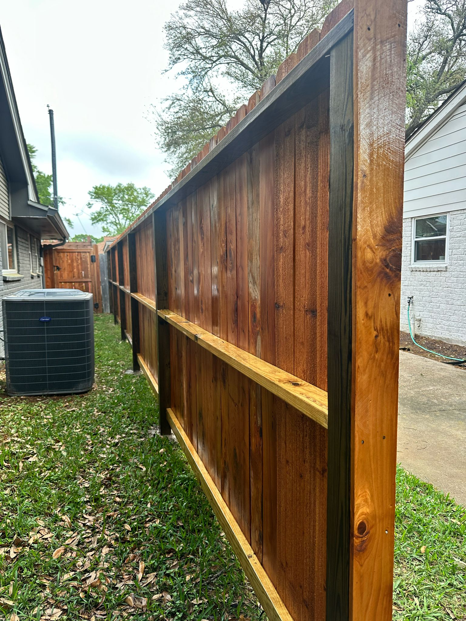 A newly installed wooden fence made of stained wood panels in a backyard, with an air conditioning unit to the left and a concrete pathway on the right.