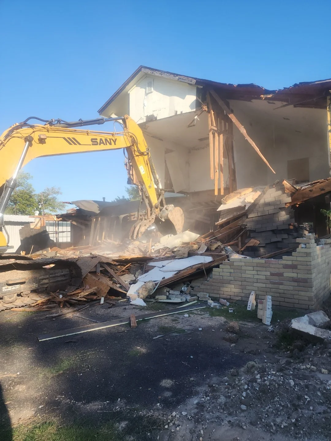 Construction excavator demolishing a two-story house, debris and dust in the air, clear blue sky.