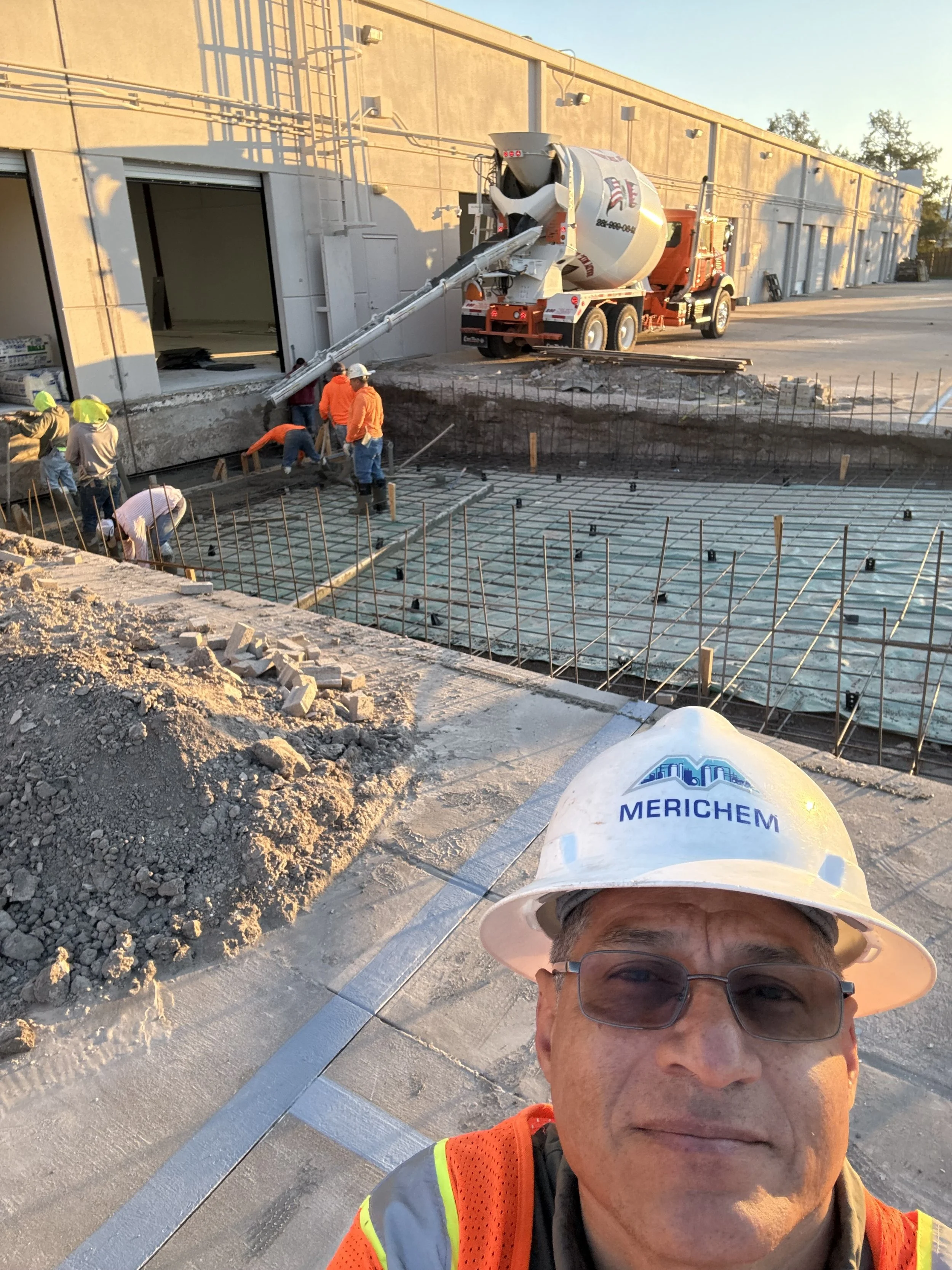 Construction site with workers pouring concrete in a foundation, a cement mixer truck in the background, and a man in a hard hat and safety vest taking a selfie in the foreground.