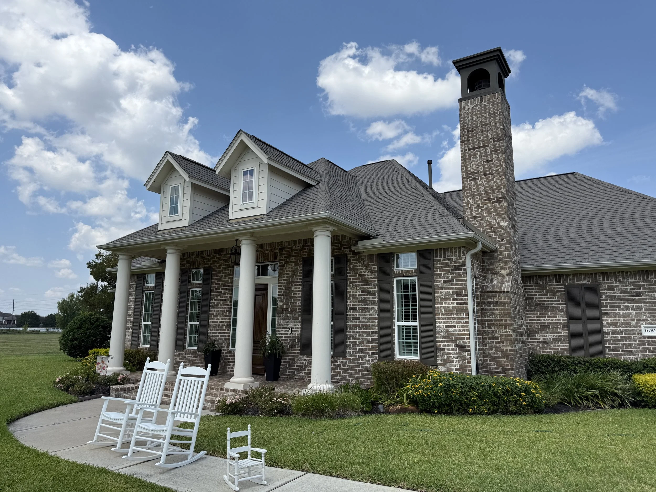 Brick house with porch, white columns, front yard with lawn, rocking chairs, and a bright sky
