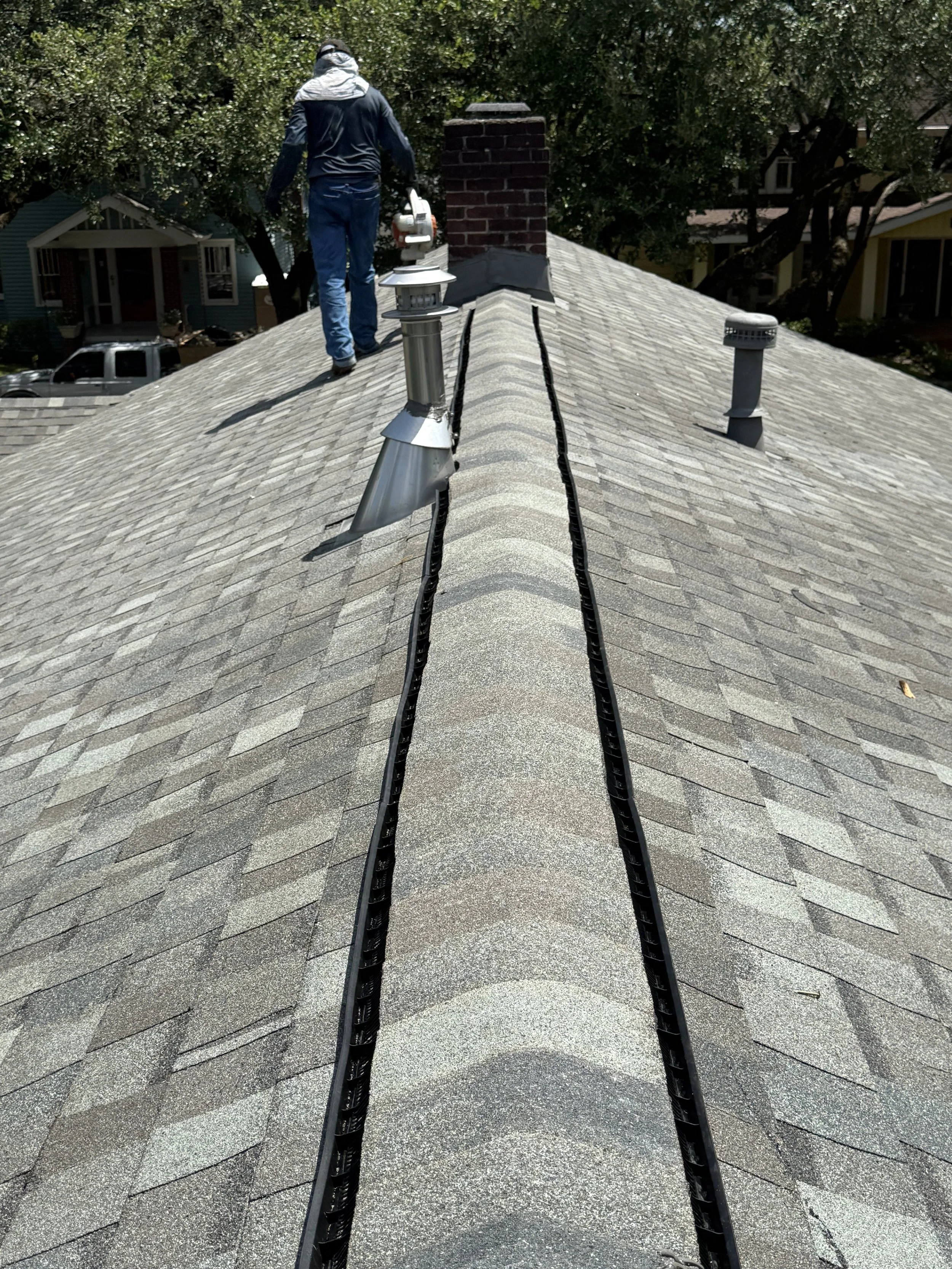 A person on a roof with shingles, using a power tool, wearing a gray hoodie and dark jacket, with trees and houses in the background.