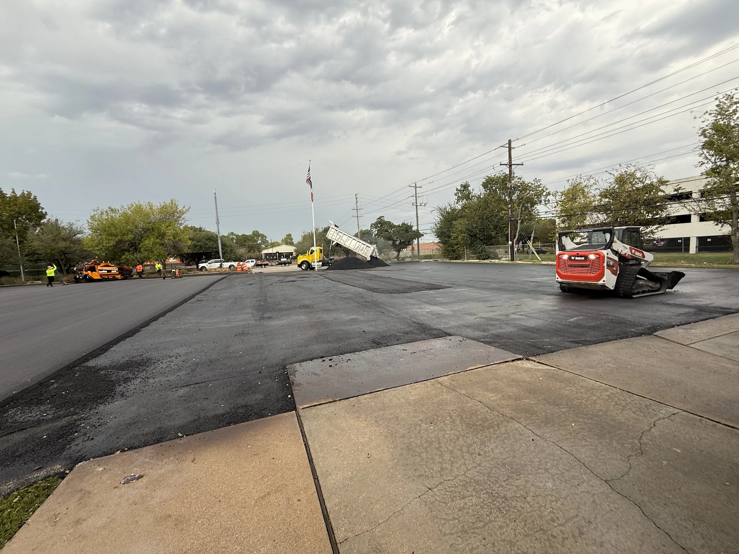 A construction site shows asphalt paving on a street with construction equipment and workers. The scene includes a dump truck, a small excavator, and workers wearing safety vests. The sky is cloudy, and there are trees and buildings in the background