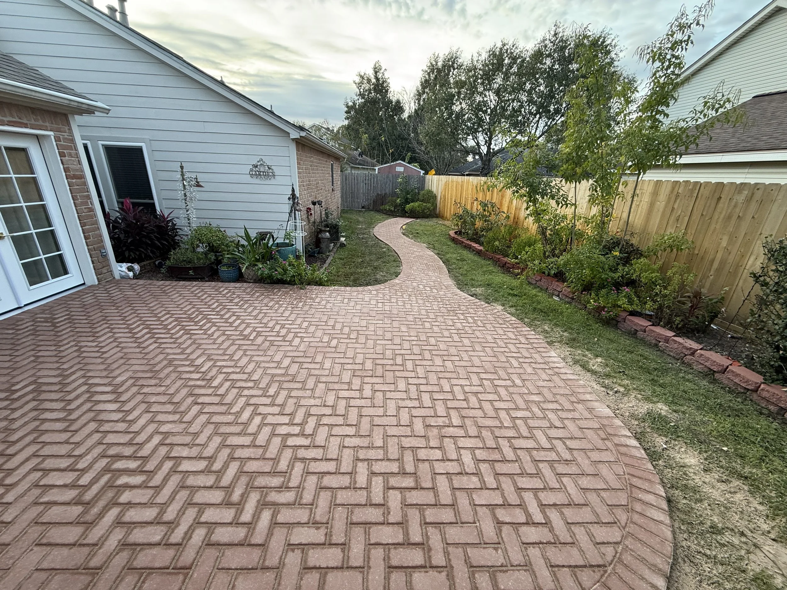 A backyard scene with a curved brick patio and a brick border garden bed on the right, containing various plants and small trees. A house with white siding and a sliding glass door is on the left, with outdoor decorations and potted plants near the h
