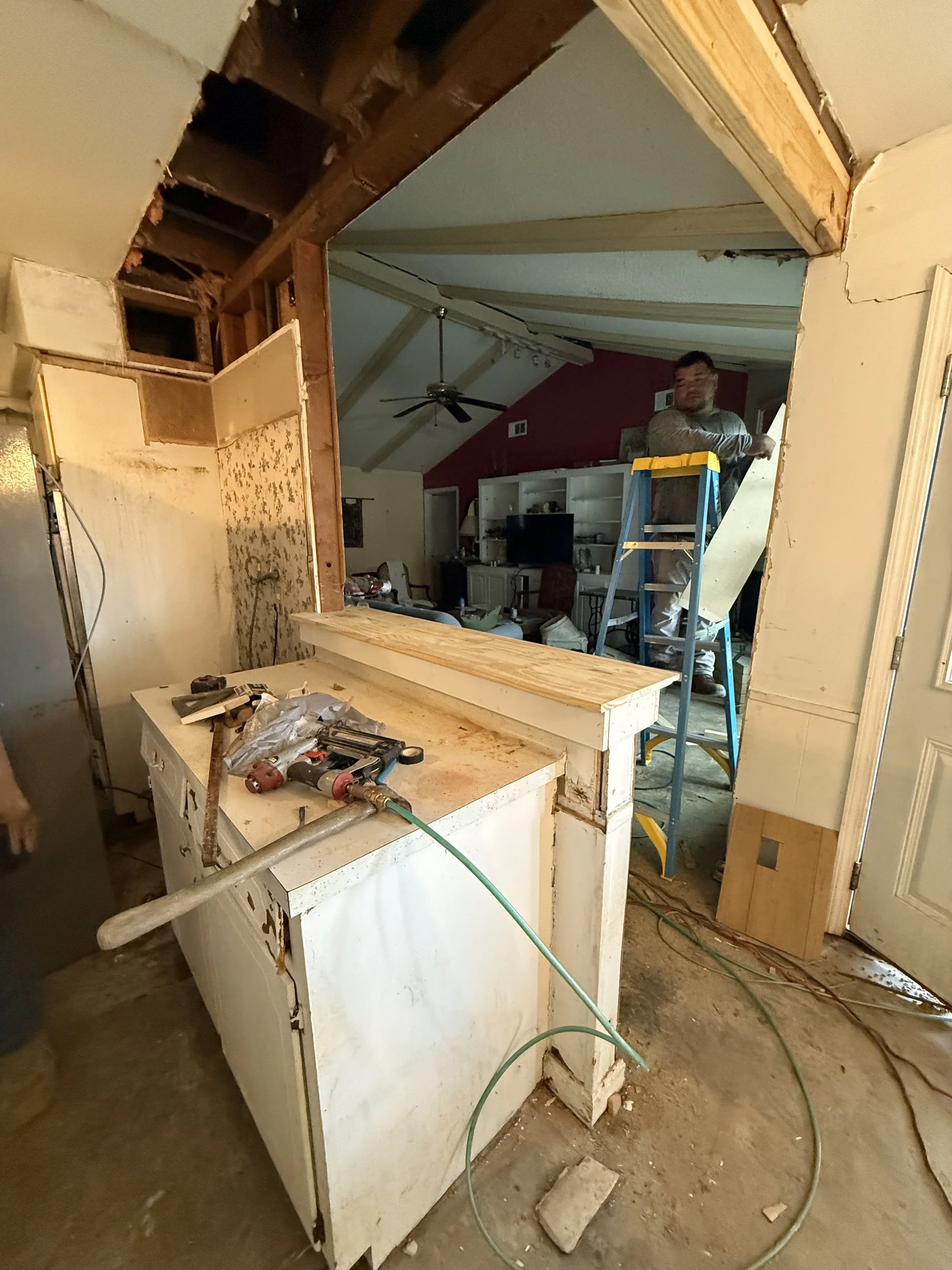 Interior of a house undergoing renovation, showing a partially demolished wall, a work table with tools, a ladder, and a person working in the background.