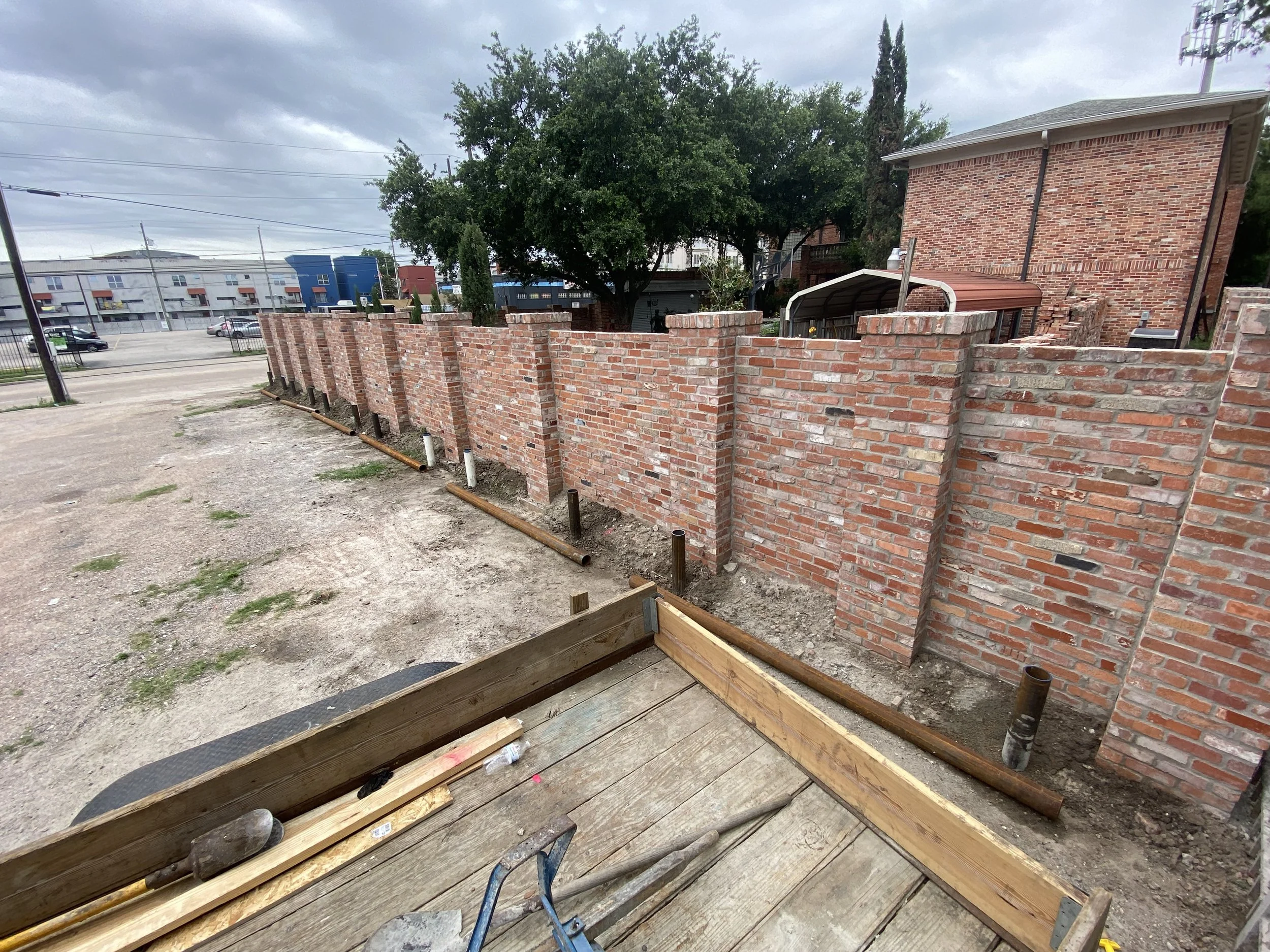 A brick wall under construction with metal pipes and dirt in front, next to a gravel parking lot with trees and buildings in the background.