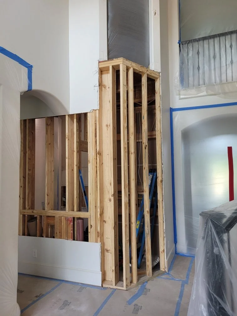 Wood framing in progress for a wall, with some sections covered in plastic and painter's tape, in a room under construction.